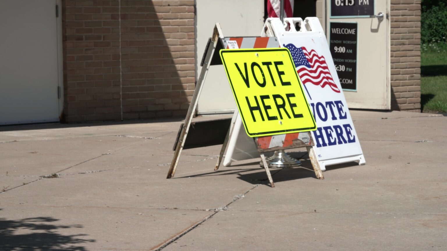 Two folding highway and sandwich board signs read Vote Here stand on a concrete patio outside the entrance to a brick building.