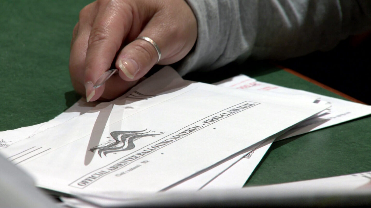 An elections worker uses a letter opener to unseal a stack of absentee ballot envelopes on top of a table.