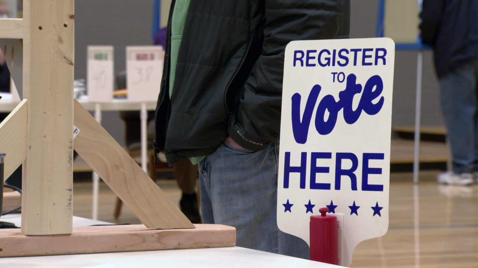 A sign reading Register to Vote Here stands next to a table with a wood-framed physical distancing divider and one person standing next to it, with tables, signs and other people standing in the background.