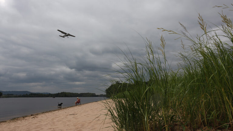 A beachgoer sits on a folding chair and a dog stands in the water of a beach as a single-engine propeller aircraft flies overhead under a cloudy sky, with marsh grasses in the foreground and tree-lined shores and bluffs in the background.