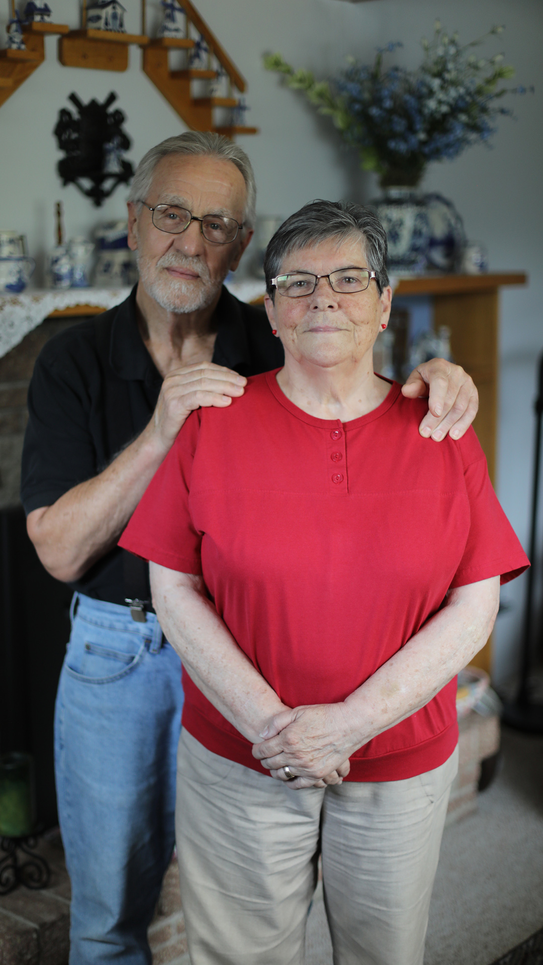 Jim Boisen and Margie Walker pose for a portrait inside a room, with displays of Delft china and a floral arrangement in the background.