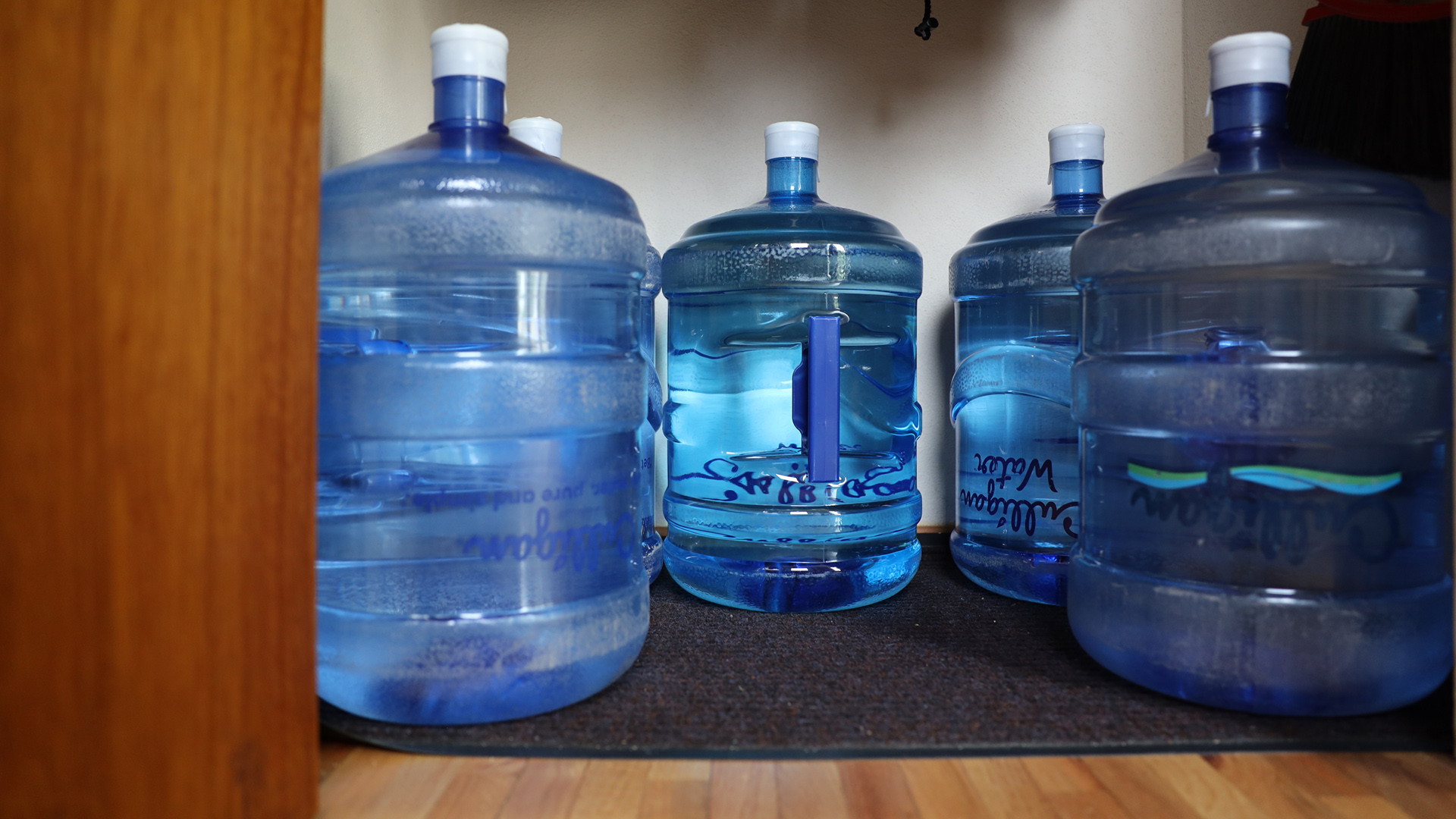 Four 5-gallon jugs of water sit at the base of a closet on a floor mat atop a wooden floor.
