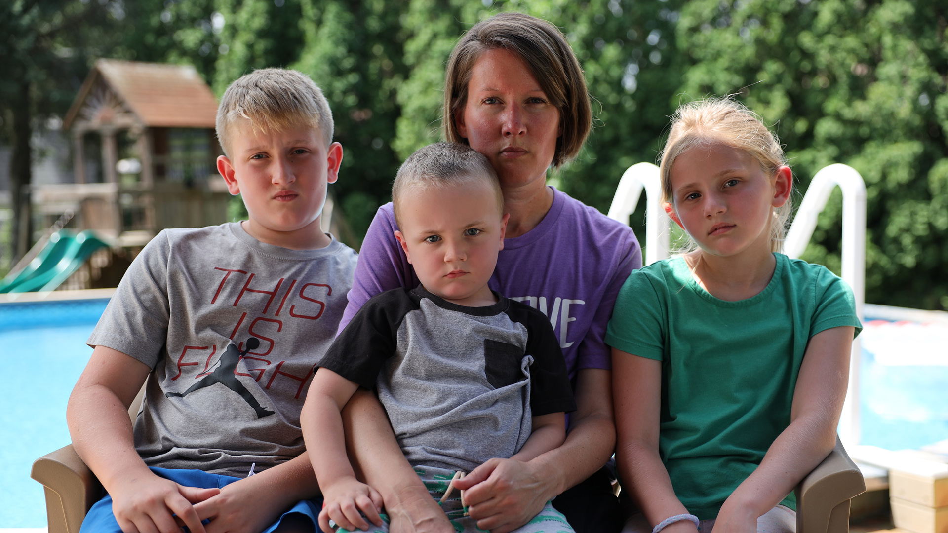 Jameson, Jackson, Heather and Ava Brice pose for a portrait while seated outdoors, with a pool, play structure and trees in the background.