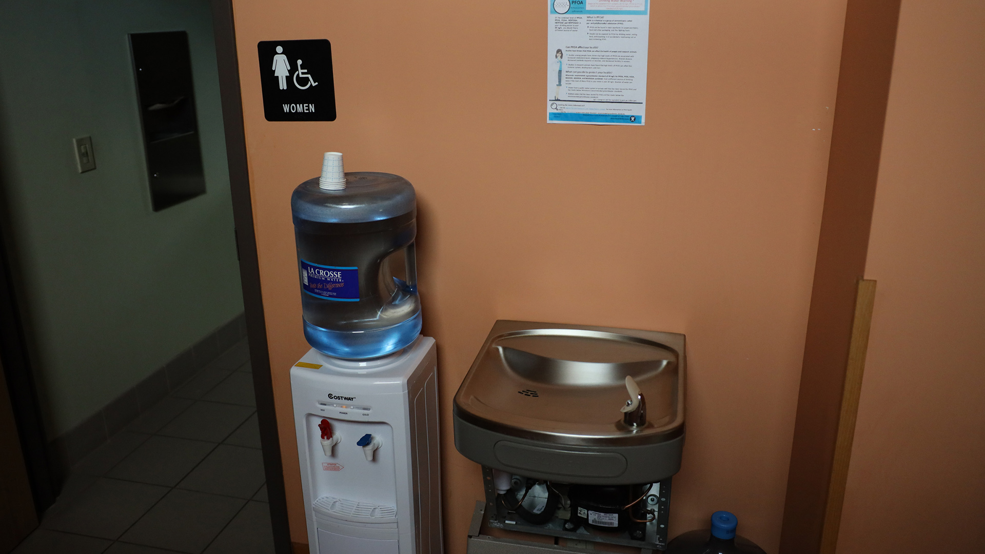A water dispenser stands next to a drinking water fountain that has been disconnected from a water connection, with a flyer warning about PFAS attached to the backing wall, adjacent to a door to a women's restroom and accompanying signage.