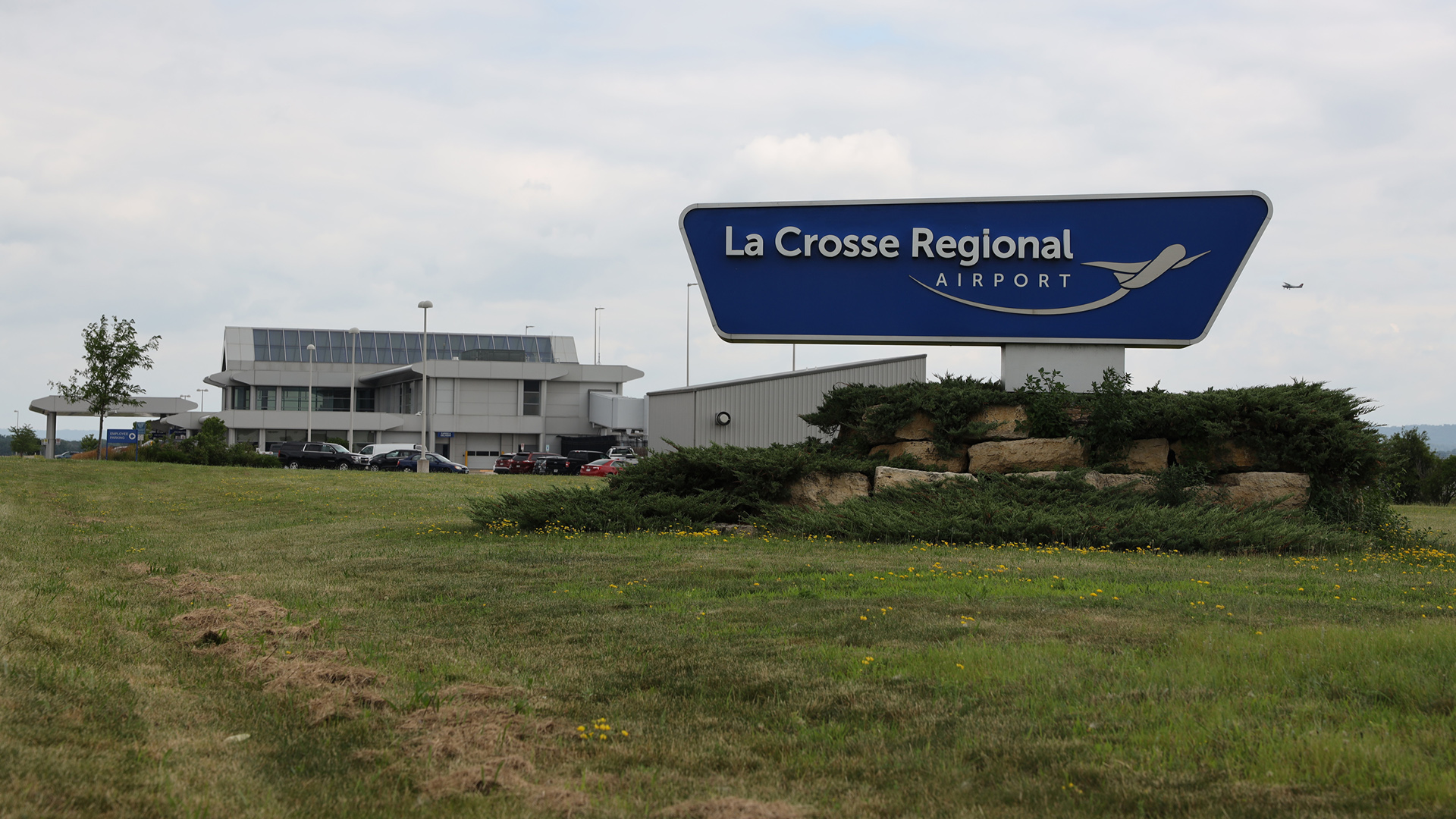 A sign reading "La Crosse Regional Airport" with a stylized graphic of a plane in flight stands in front of a multi-story building and parking lot, with a grass lawn in the foreground and a small aircraft in the sky in the background.
