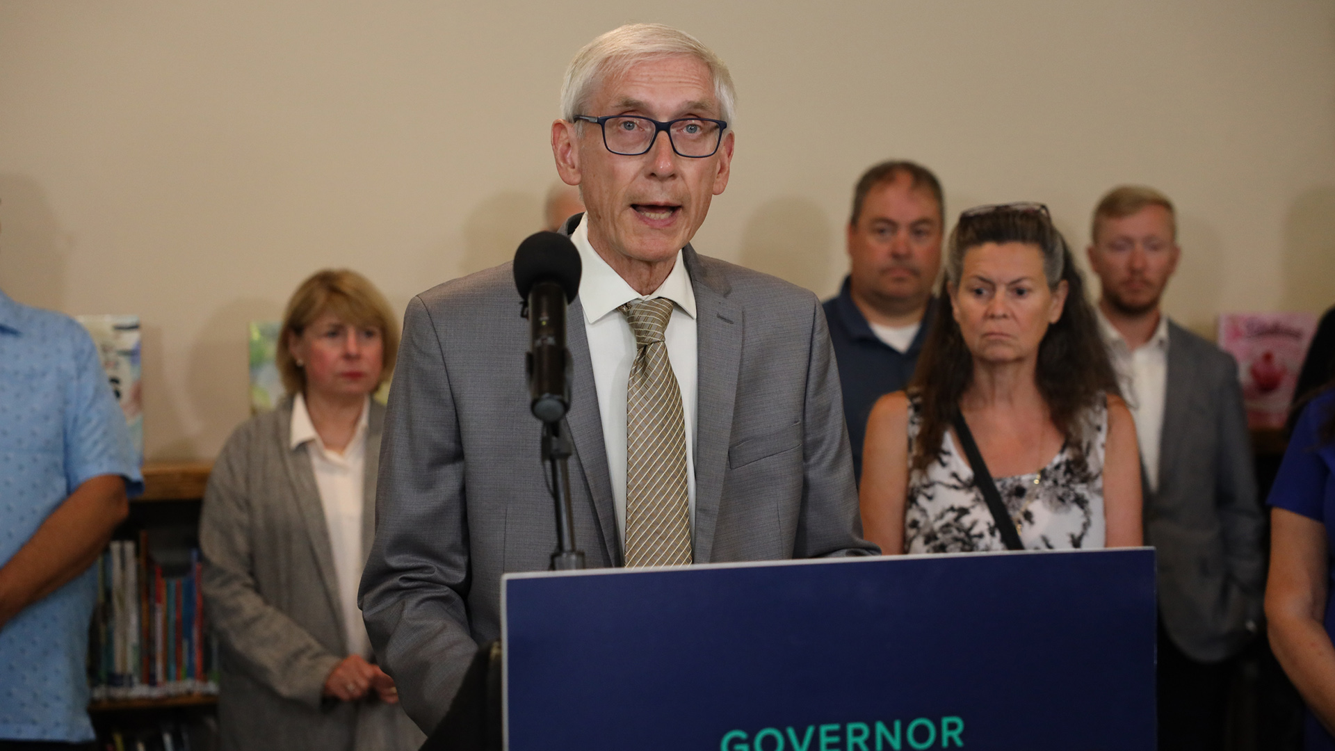 Tony Evers stands in front of a podium with one microphone and speaks with other people standing in the background.
