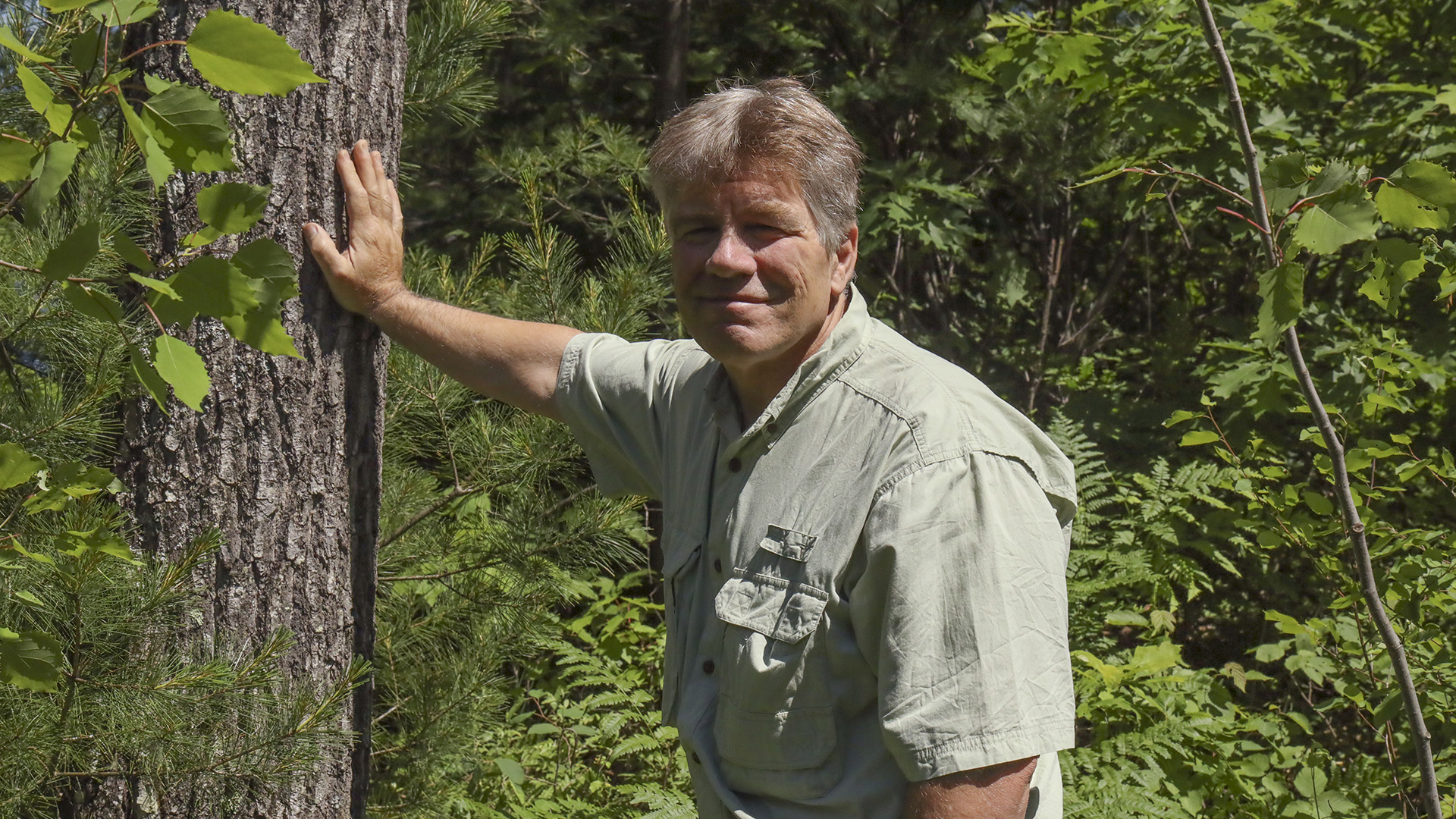 John Schwarzmann holds his right hand against the trunk of a tree with foliage in the background.
