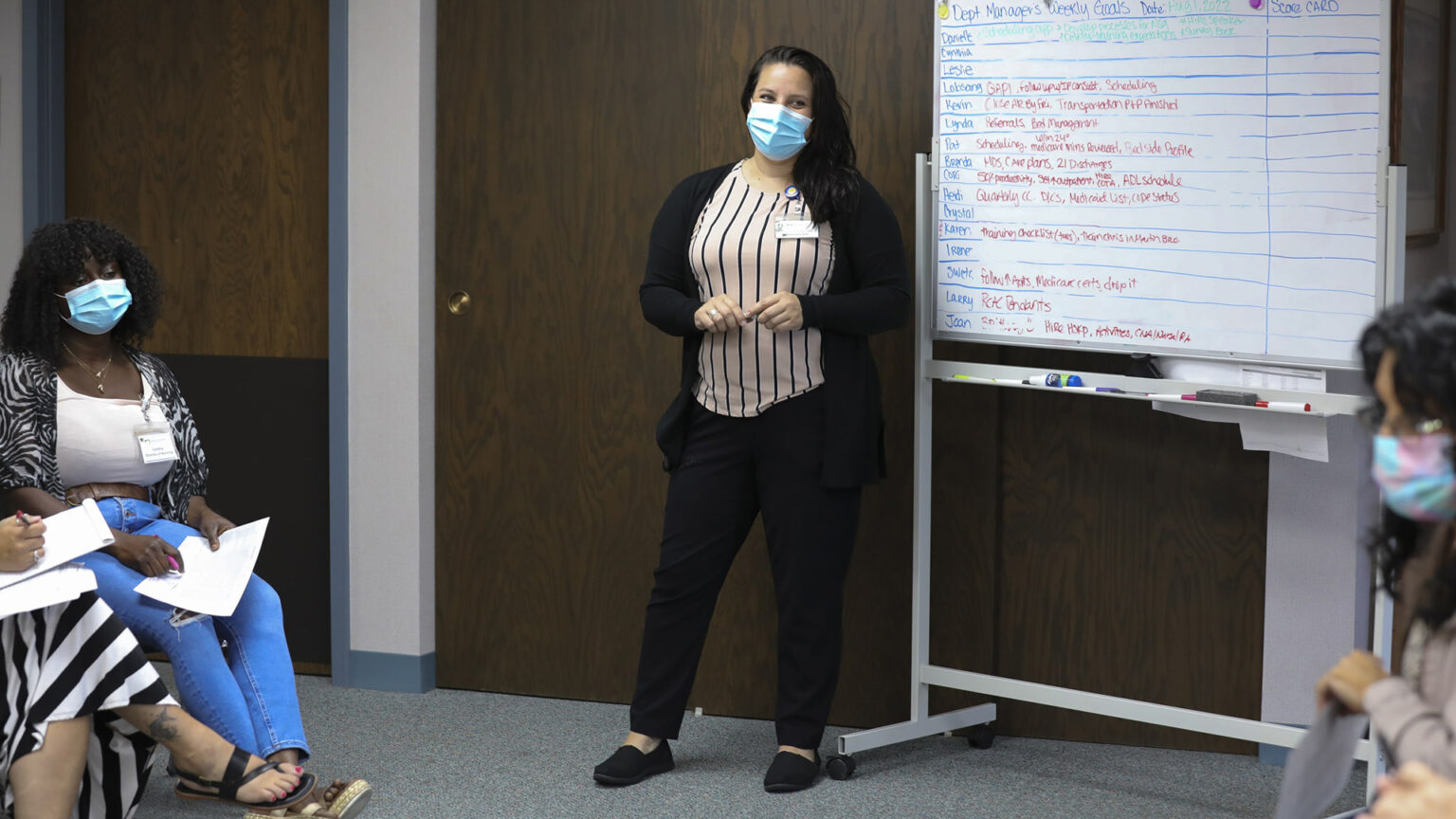 Danielle Sigler stands next to a whiteboard listing staff responsibilities, with colleagues seated on other side and wood-paneled doors in the background.