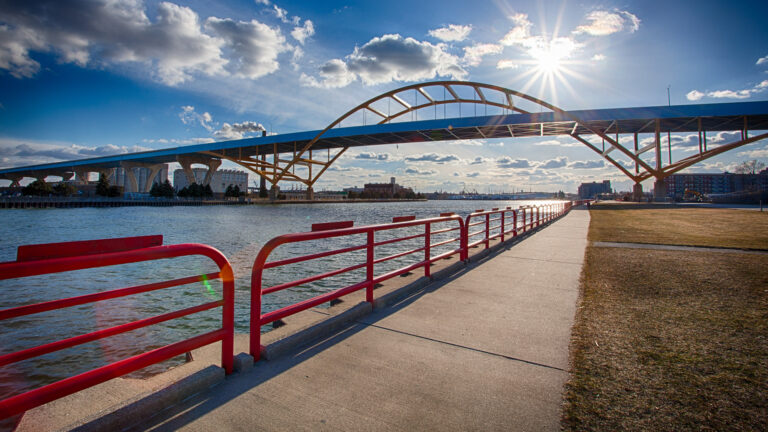 A setting sun in a partly cloudy sky shines over the Daniel Hoan Bridge, with a riverside safety railing, sidewalk and lawn in the foreground and buildings in the background.