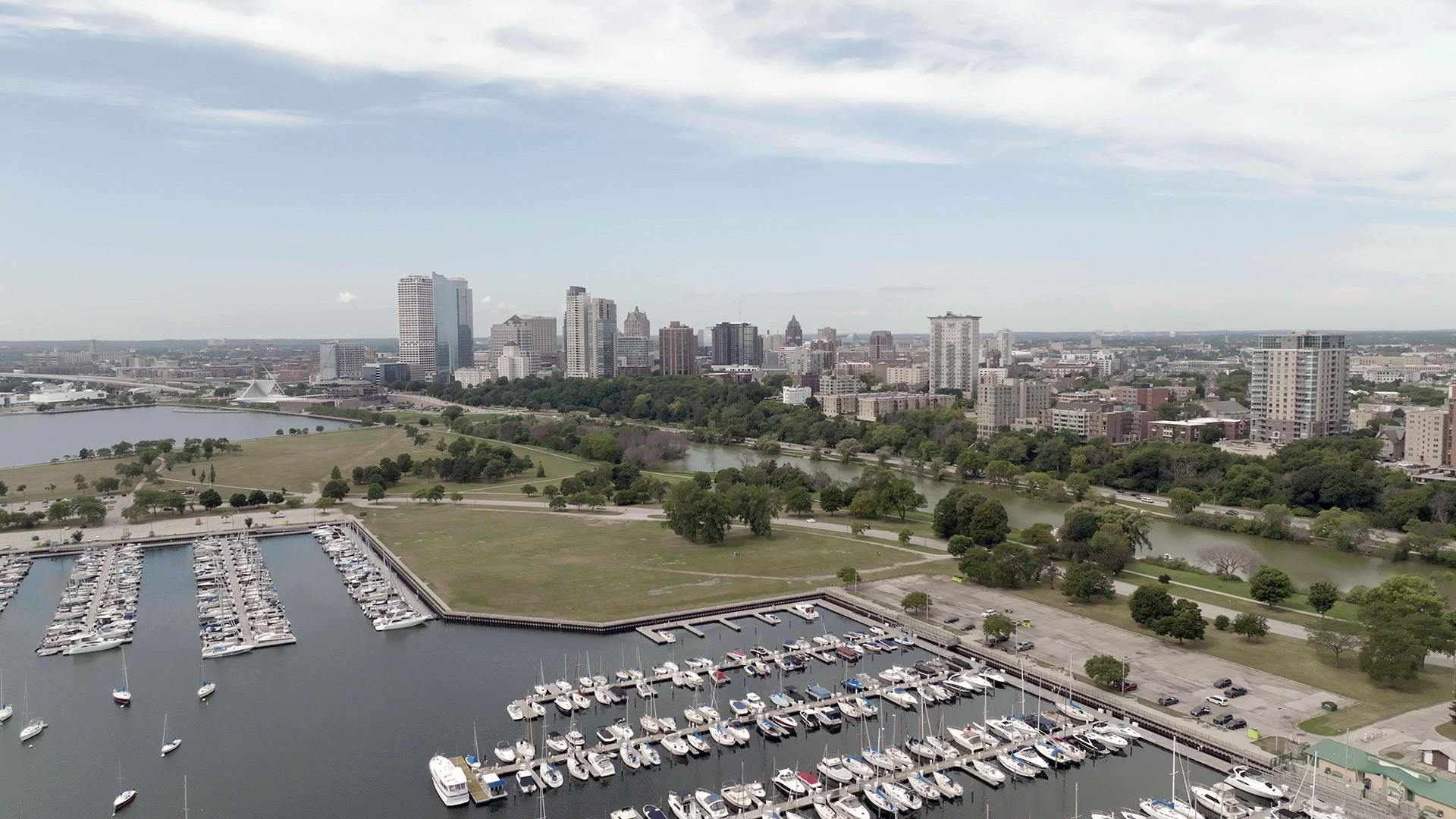 An aerial photo shows a marina filled with boats on a lakeshore sits next to a public park with numerous trees in front of an urban skyline with tall buildings under a partly-cloudy sky.