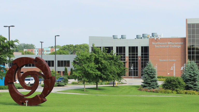 An large abstract metal sculpture in the form of a wheel stands on a lawn in front of a multi-section and multi-story building with brick, metal and glass walls with signs that read Northeast Wisconsin Technical College and Student Center with trees in the foreground and background.