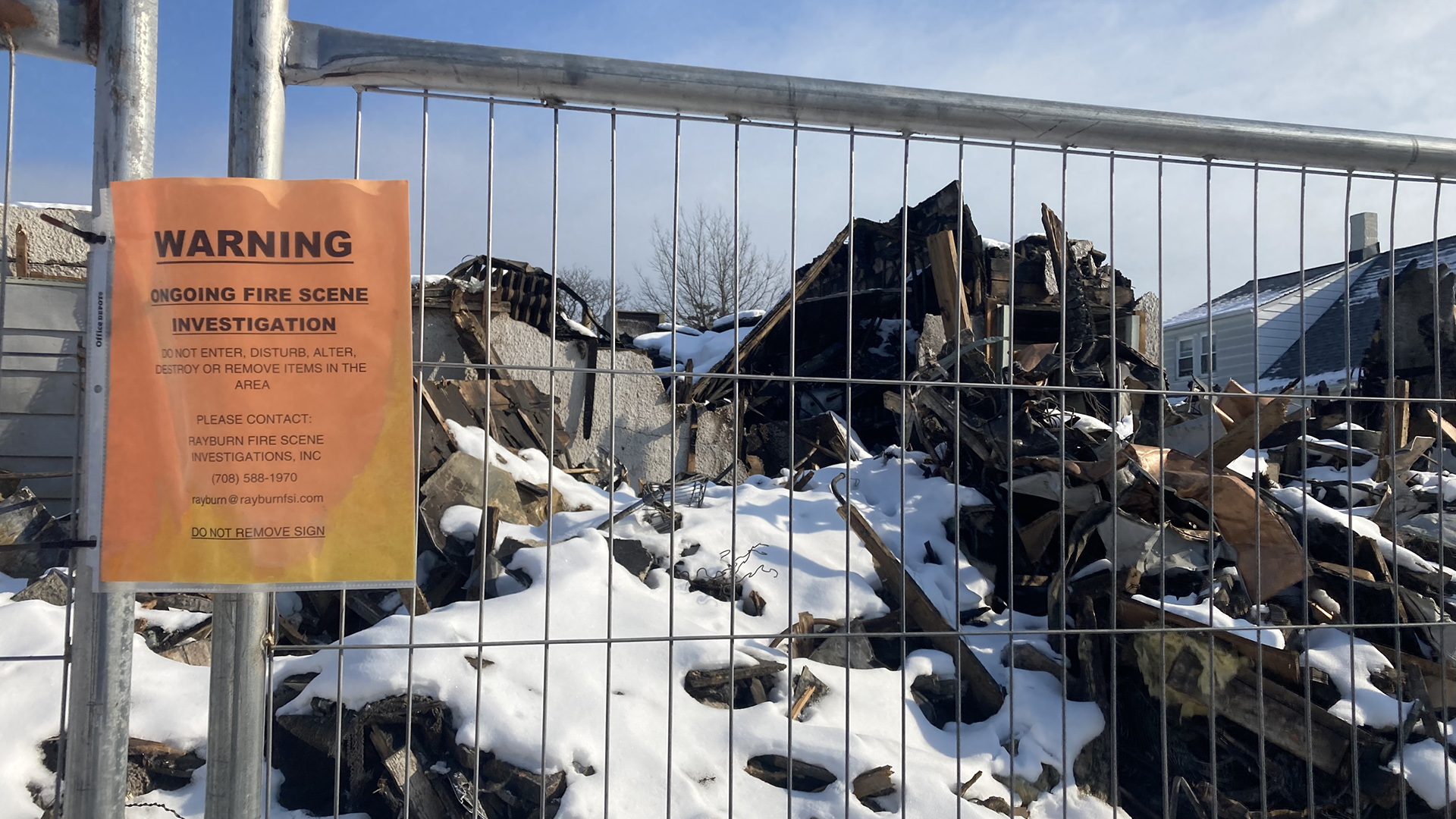 A paper sign that reads "Warning" and "Ongoing Fire Scene Investigation" is posted to a metal fence in front of a snow-covered pile of charred rubble, with other buildings standing in the background.