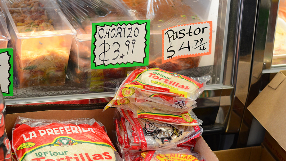 Stacks of packaged flour tortillas stand in front of the window of a refrigerated counter with displays and prices for chorizo and pastor.