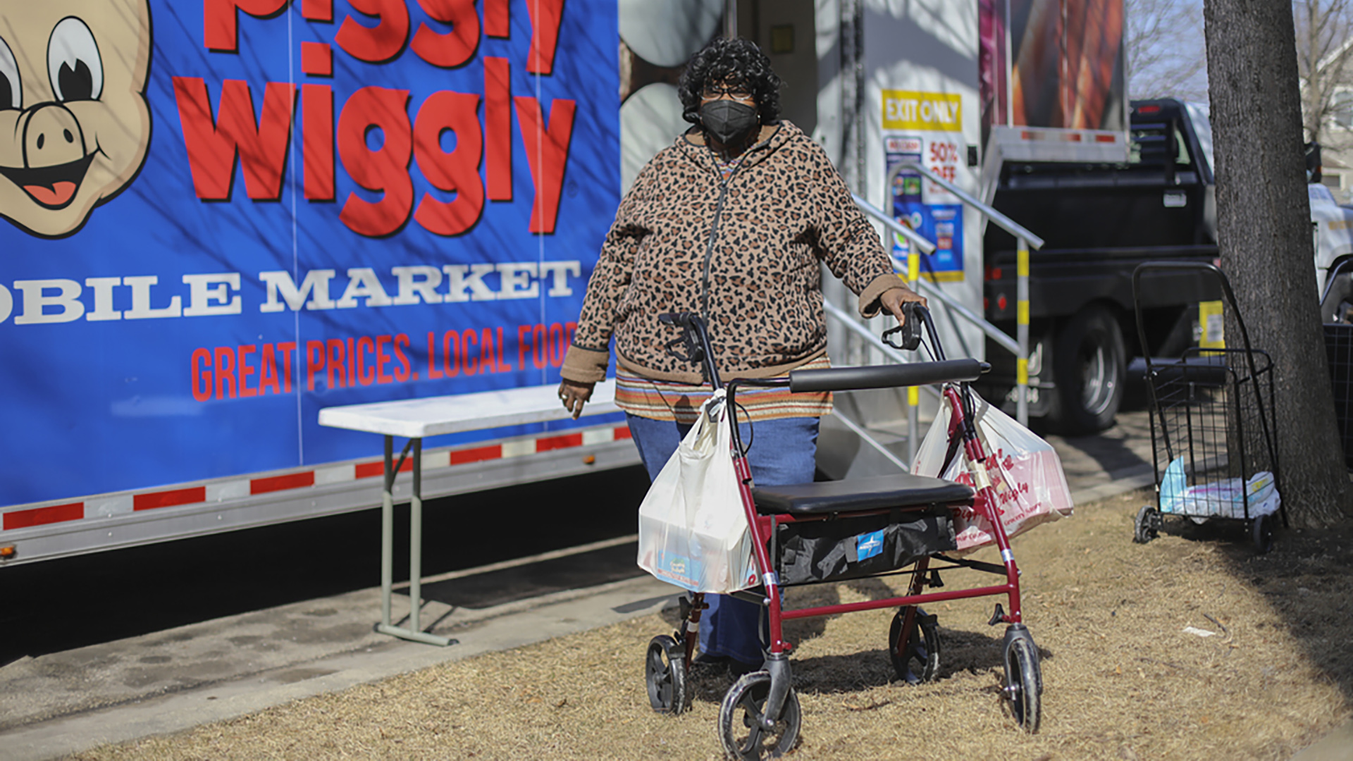 Shirley Johnson stands behind a mobility walker with two filled plastic grocery bags hanging on it while standing in front of a mobile market with the Piggly Wiggly logo, wordmark and a slogan reading "Great Prices. Local Food."