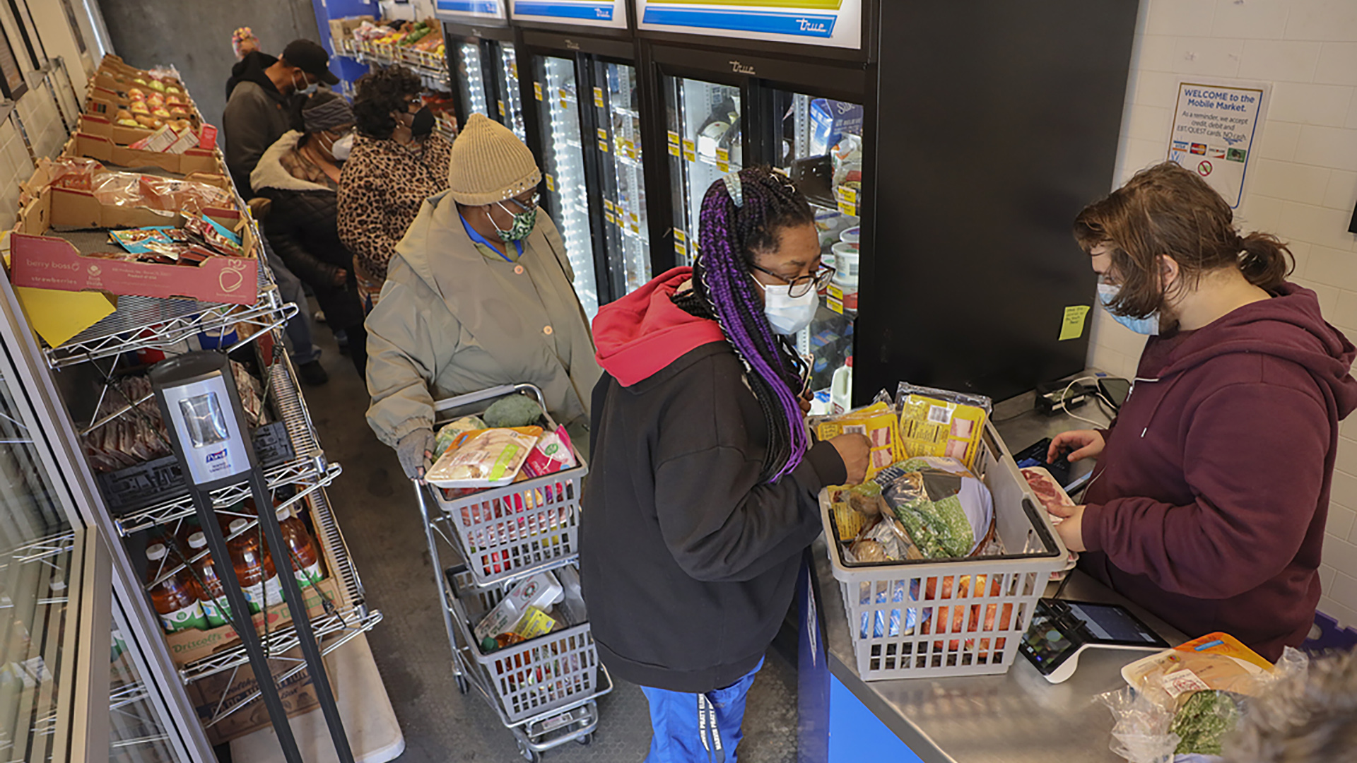 Shoppers with carts and baskets shop, select items and check out while standing in a line in a mobile food market, with shelves and coolers containing groceries on either side.