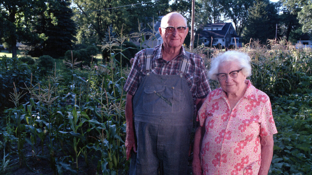 An 80s image of an old farmer in overalls standing next to his wife