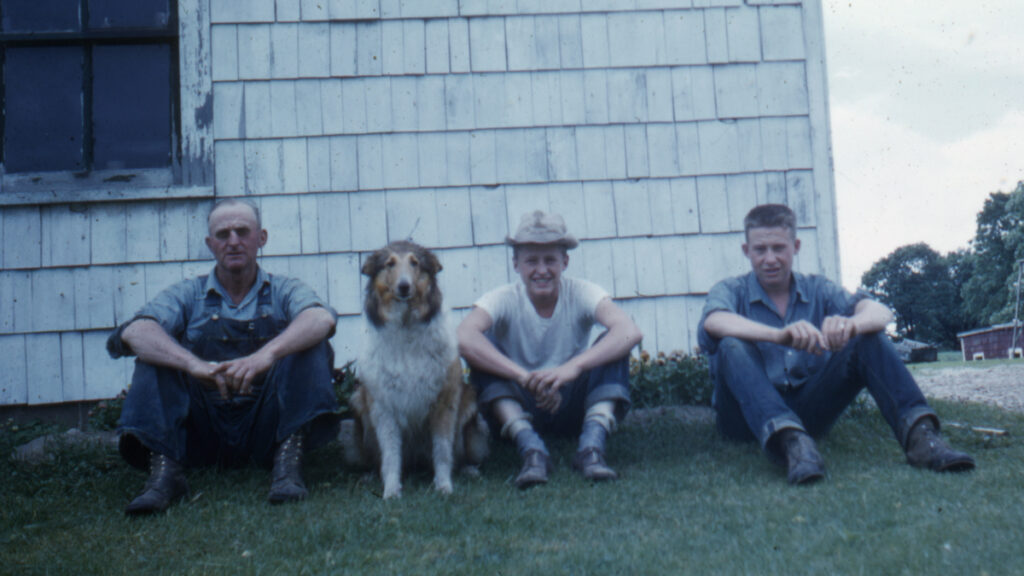 A retro image of a farmer, his two sons and a collie smiling as they sit for a picture outside
