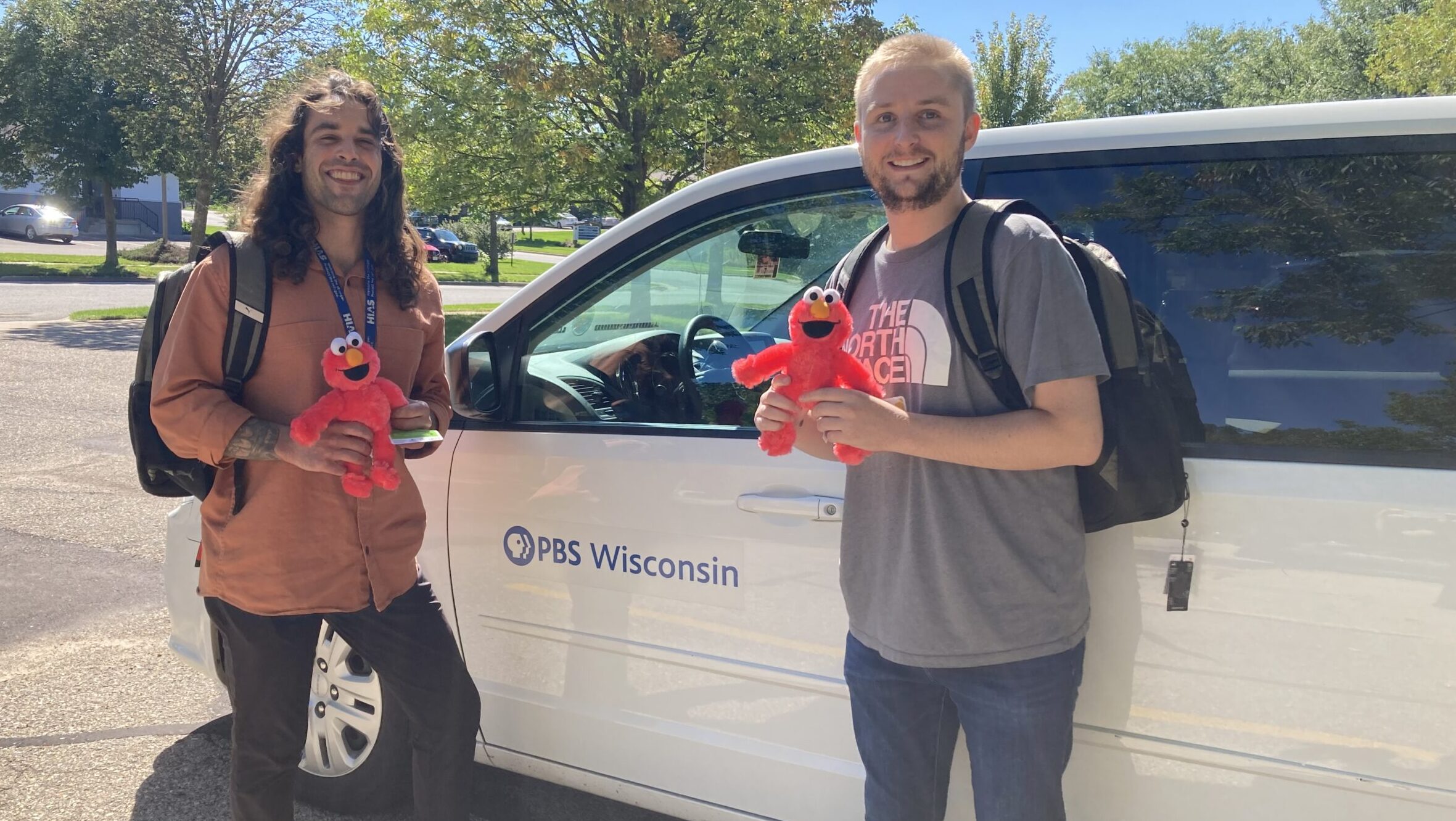 two men holding Elmo stuffed animal, standing by PBS Wisconsin van