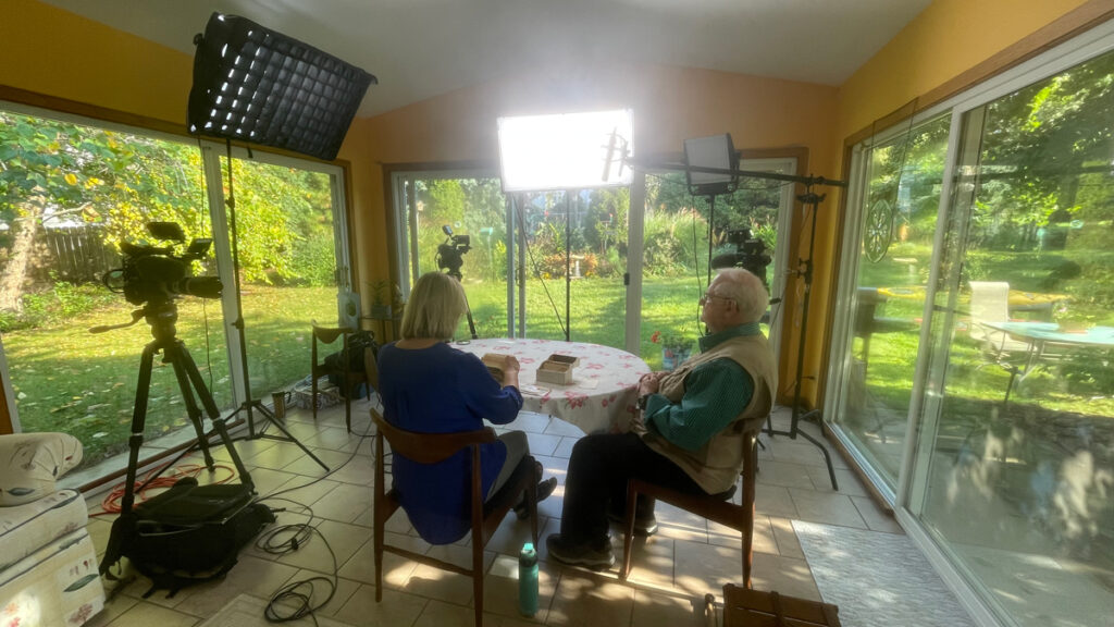Susie Apps-Bodilly and Jerry Apps sit in her sun porch for an interview. Their backs are to the viewer and lighting equipment is in front of them. 