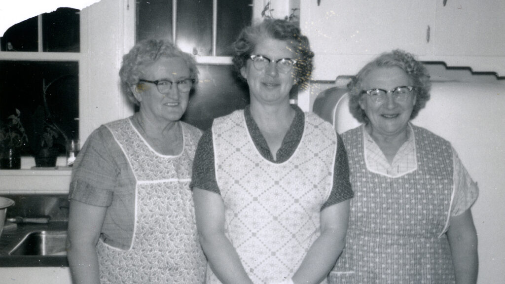 Three older sisters in aprons pose in a black and white picture of a farm kitchen