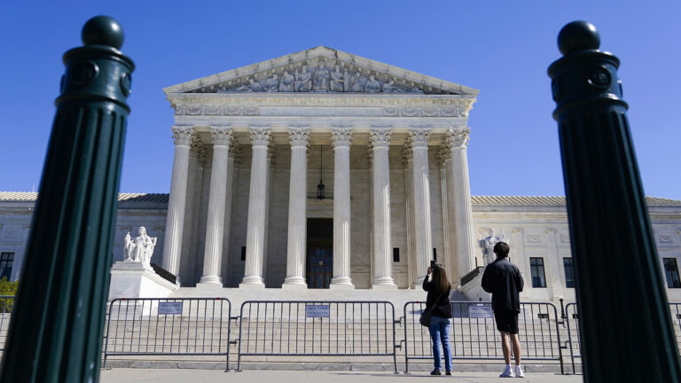 Two people stand behind metal crowd barriers at the base of steps leading to the front façade of the U.S. Supreme Court Building with its portico of columns, framed by two metal parking bollards in the foreground.