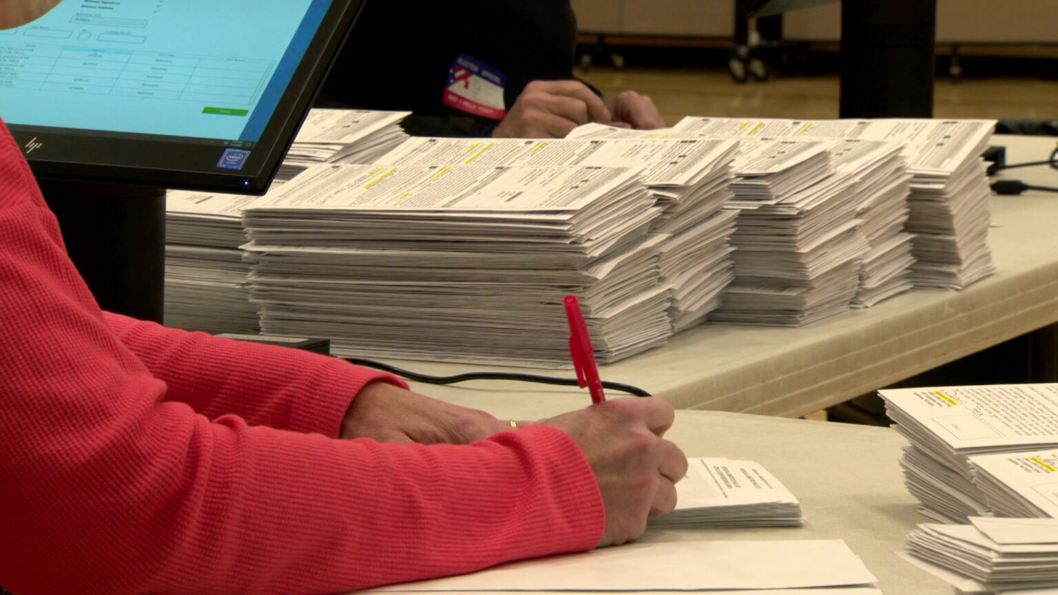 The arms of a seated person use a pen to mark an absentee ballot envelop on the surface of a folding table, with stacks of additional envelopes on the tabletop and another election worker conducting the same work at an adjacent table with multiple stacks of envelopes.
