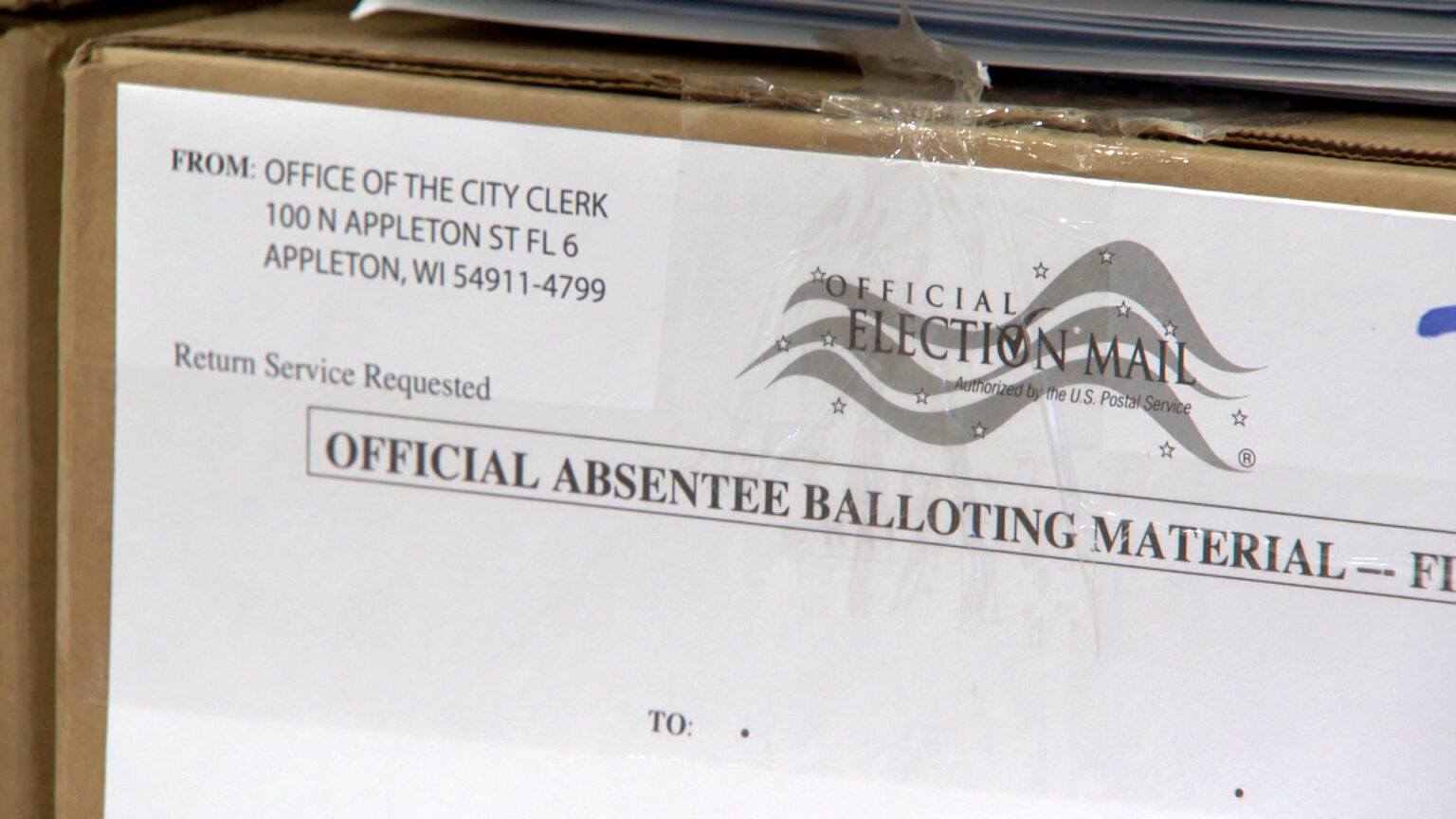 An label taped to a cardboard box reads Official Absentee Balloting Material with a return address for the city clerk of Appleton.