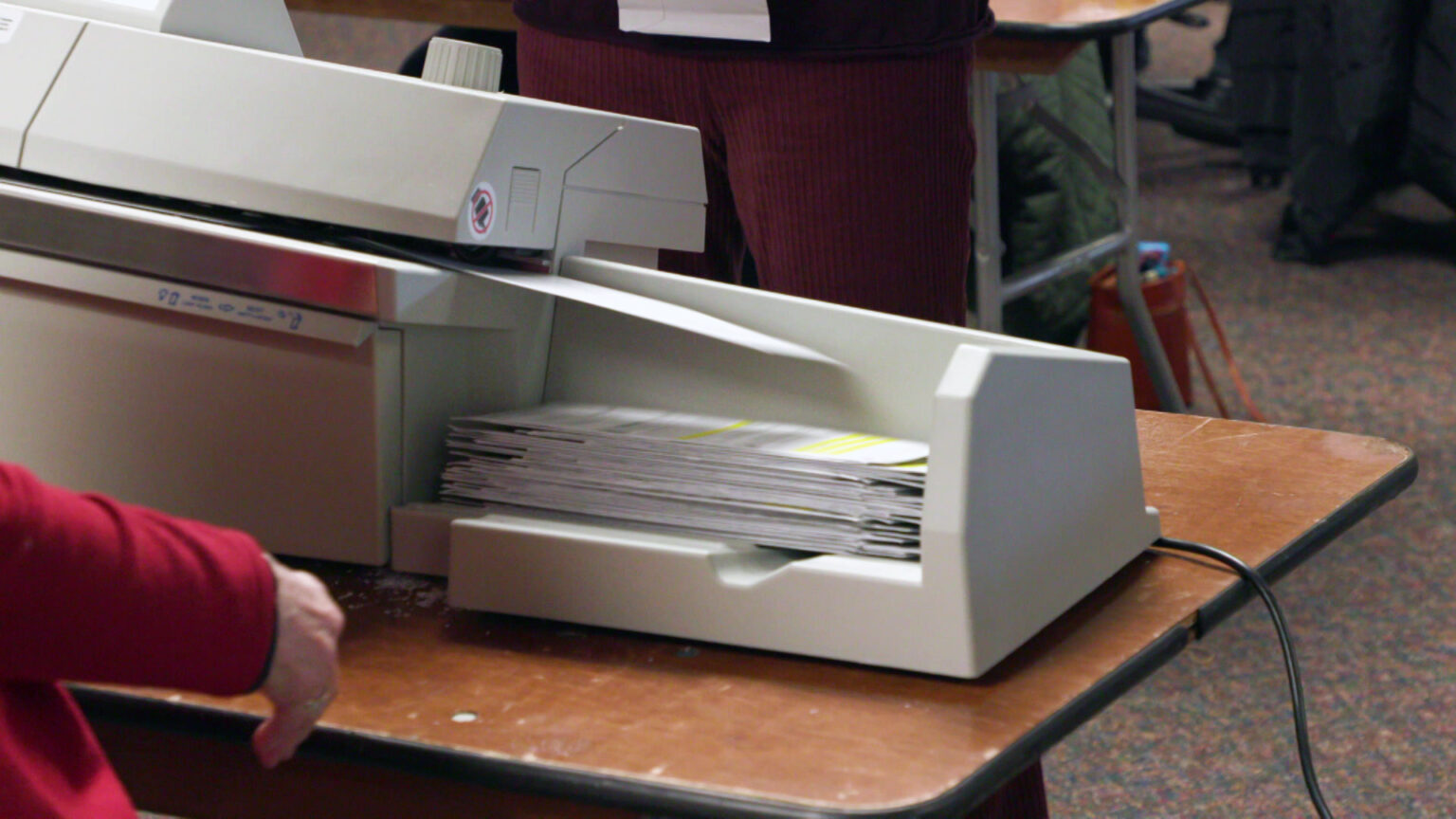 Absentee ballot envelopes are processed by an envelope-opening machine set on a folding table in a carpeted room.