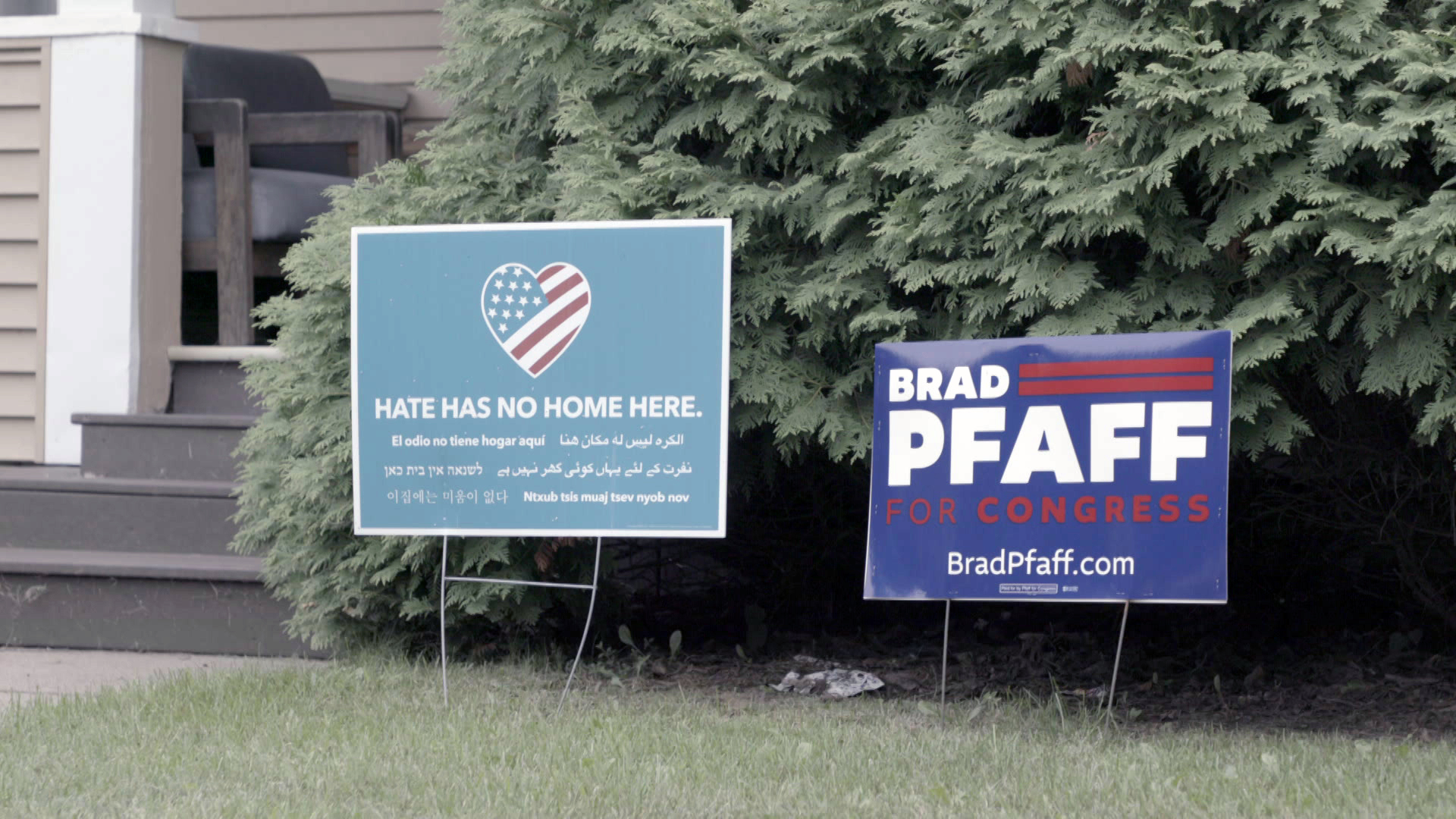Two yard signs are placed in a lawn in front of a tree and stairs leading to a patio — one reads "Hate Has No Home Here" under a U.S. flag superimposed on a heart symbol, and another reads "Brad Pfaff for Congress."