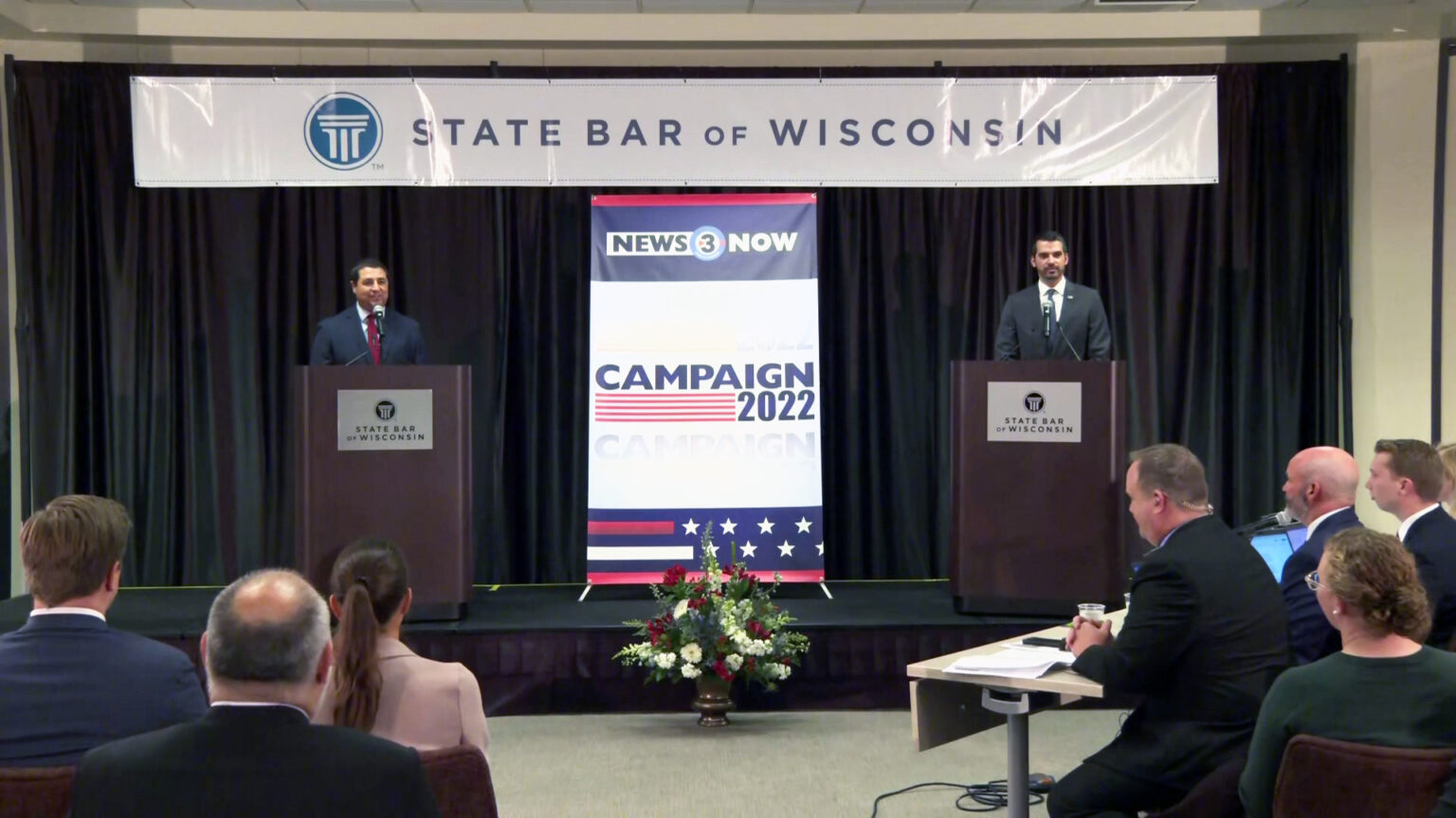 Josh Kaul and Eric Toney stand behind podiums and in front of a curtain on a low stage, with a banner above reading State Bar of Wisconsin and a display between them reading News3Now and Campaign 2022, with three people seated at a table facing stage left and other audience members seated in chairs.