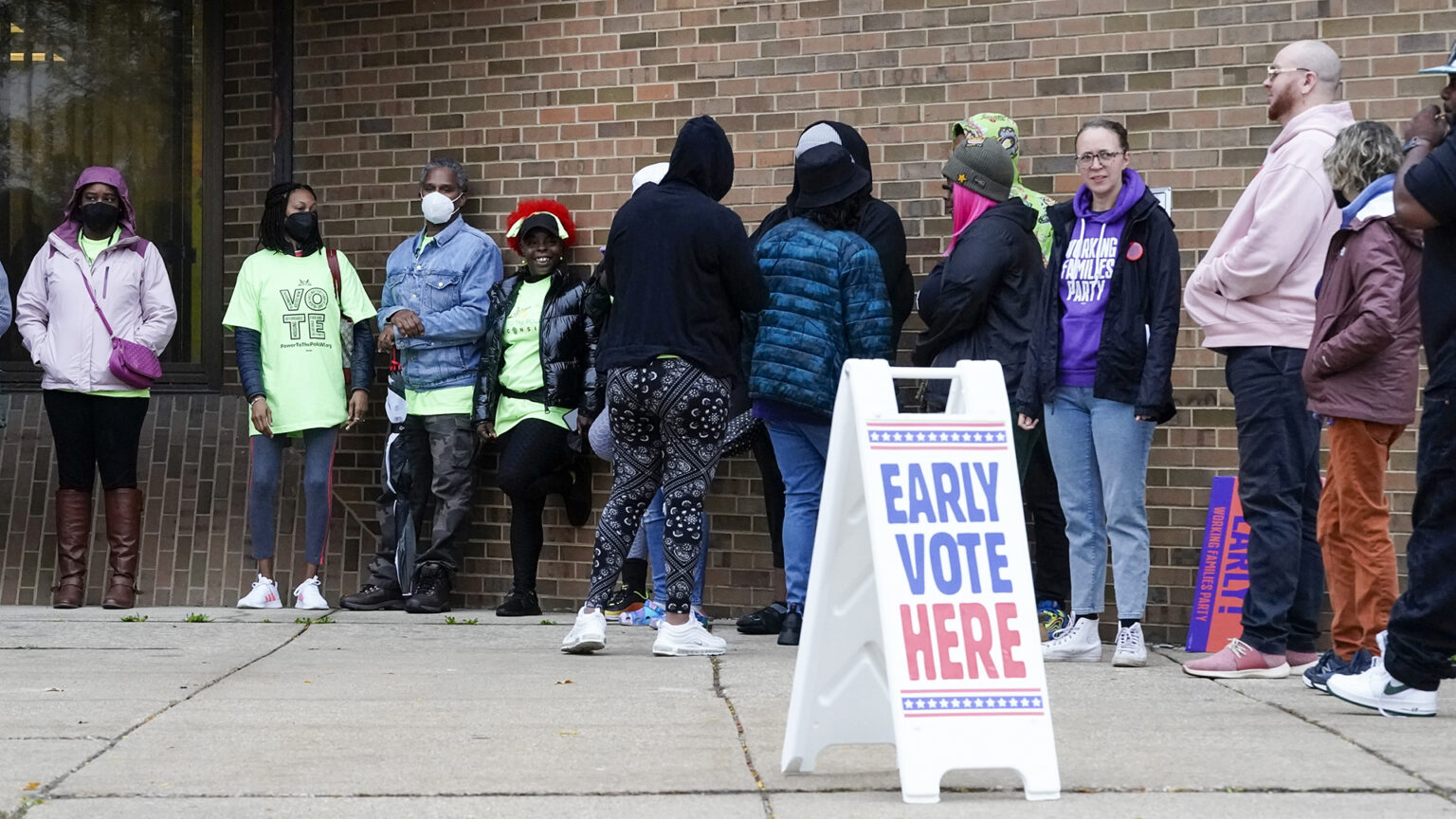 Voters stand in a line on pavement next to a brick wall, with a sandwich board sign in the foreground reading Early Vote Here.