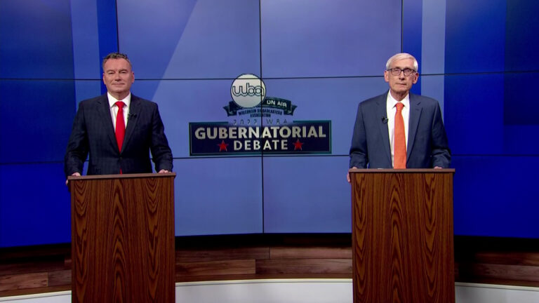 Tim Michels and Tony Evers stand behind wooden podiums on a stage set.