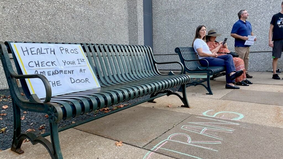 A sign propped on the back of an outdoor bench reads "Health Pros Check Your 1st Amendment at the Door," with seated and standing people and a building wall in the background.