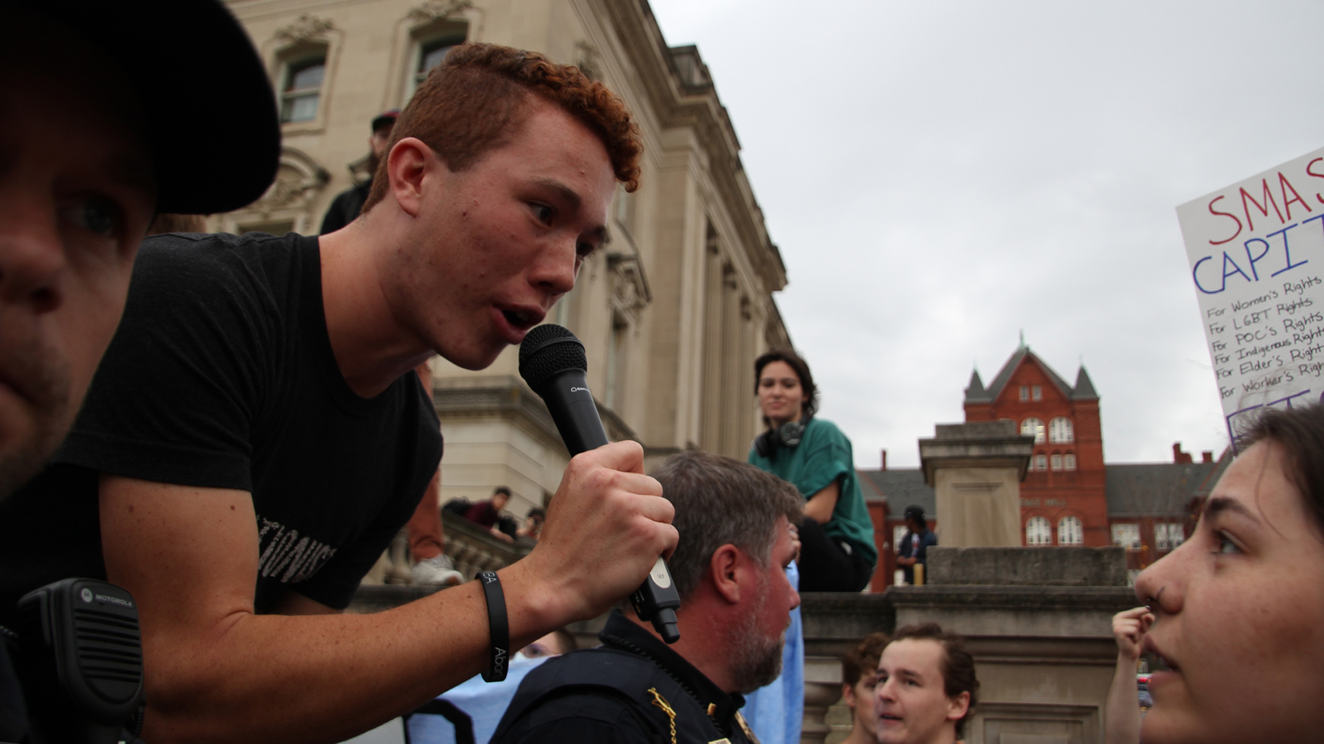 An evangelist speaking into a microphone outdoors faces and lean towards a protestor, with other people and buildings in the background.