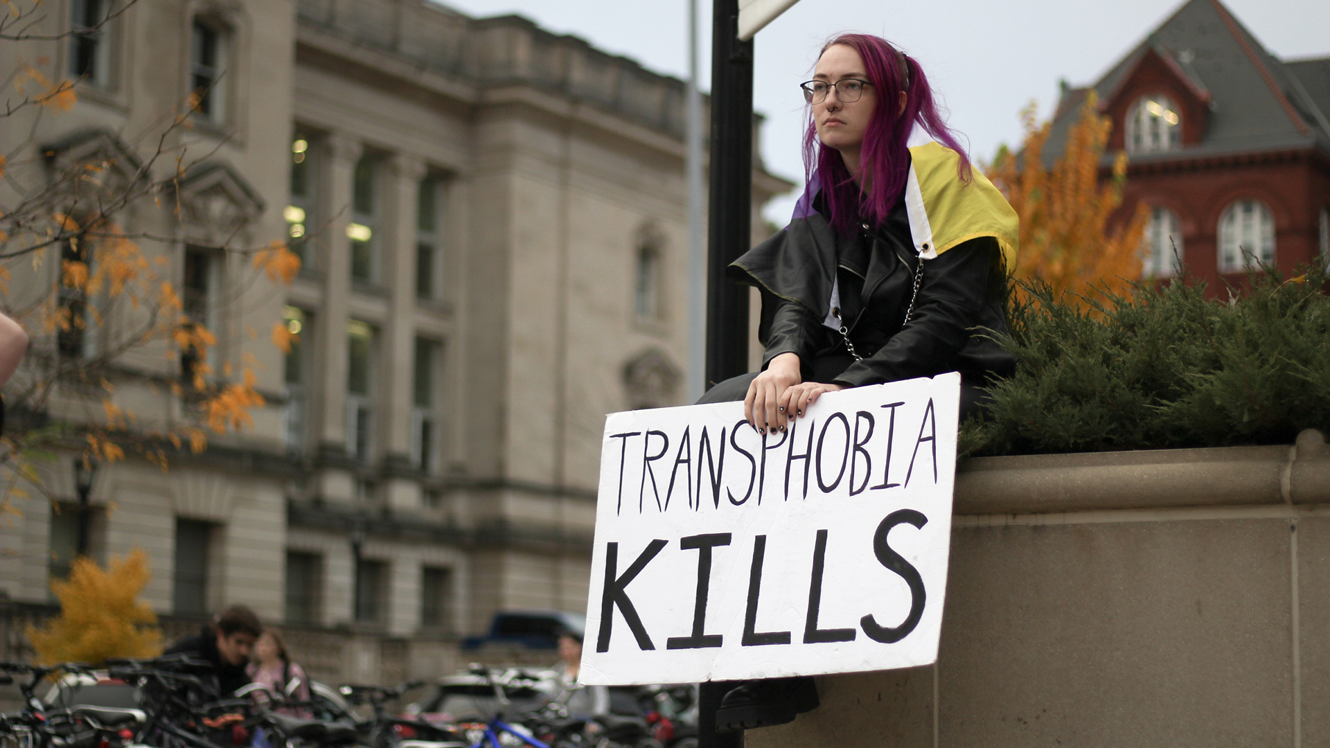 A protestor sits on the edge of a planter and holds a sign that reads "Transphobia Kills" with parked bicycles and buildings in the background.