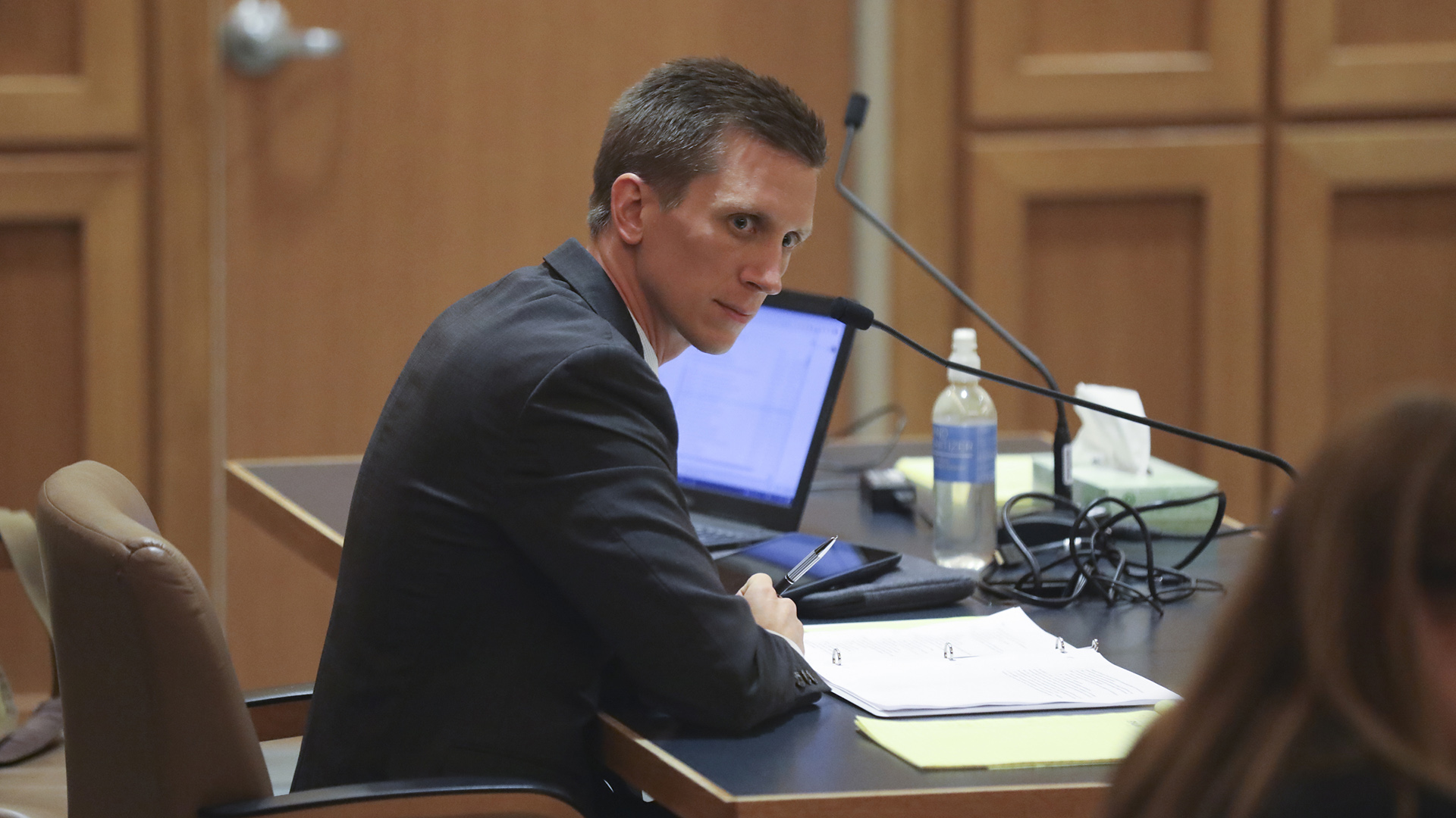 Luke Berg looks to the right while sitting at a courtroom counsel table with a three-ring binder, laptop computer, microphones, bottle of water, tissue box and other items on its surface.