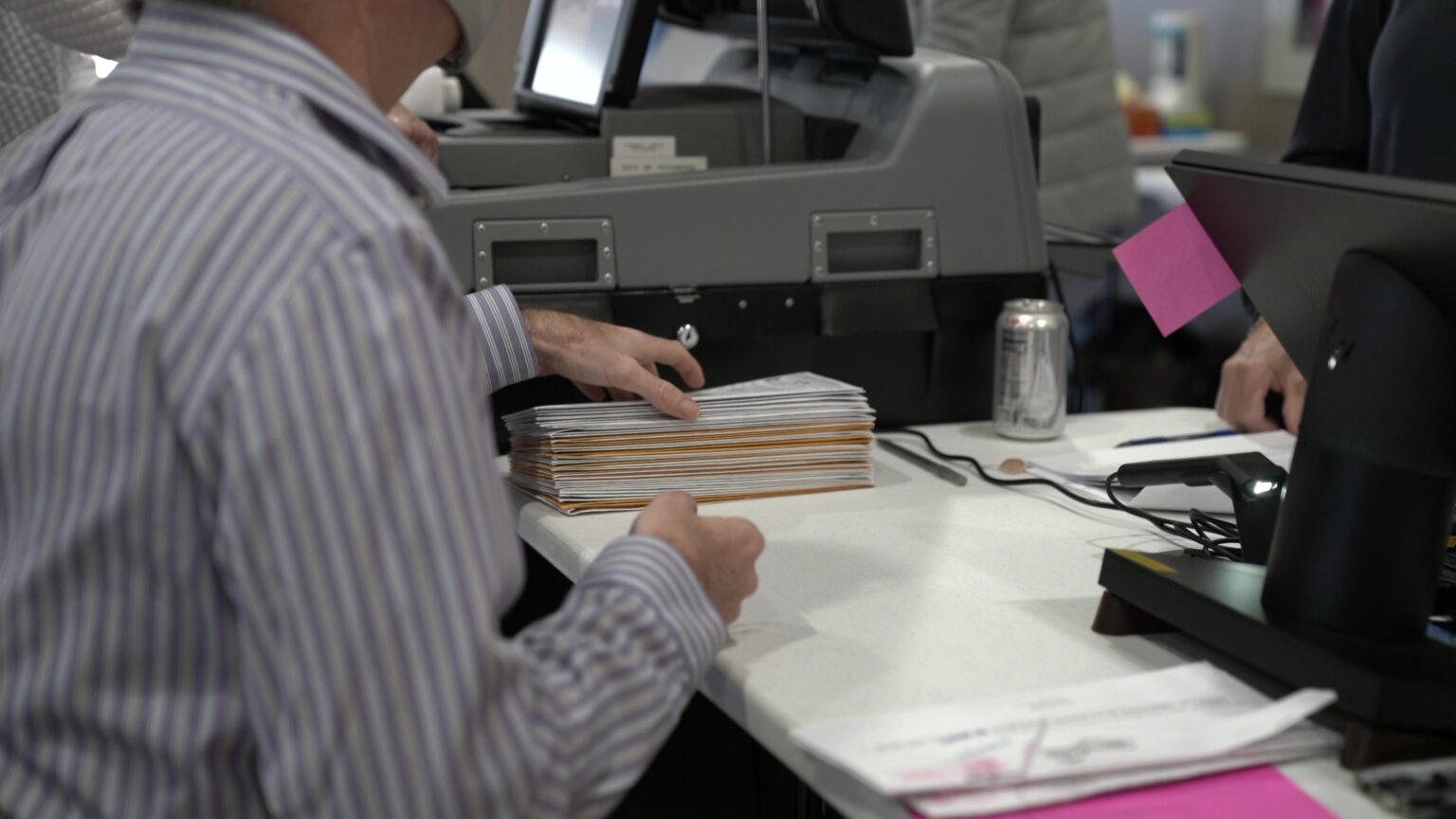 A seated person selects a folded absentee ballot envelope from the top of a stack on a table, with a ballot tabulation machine and standing people in the background.