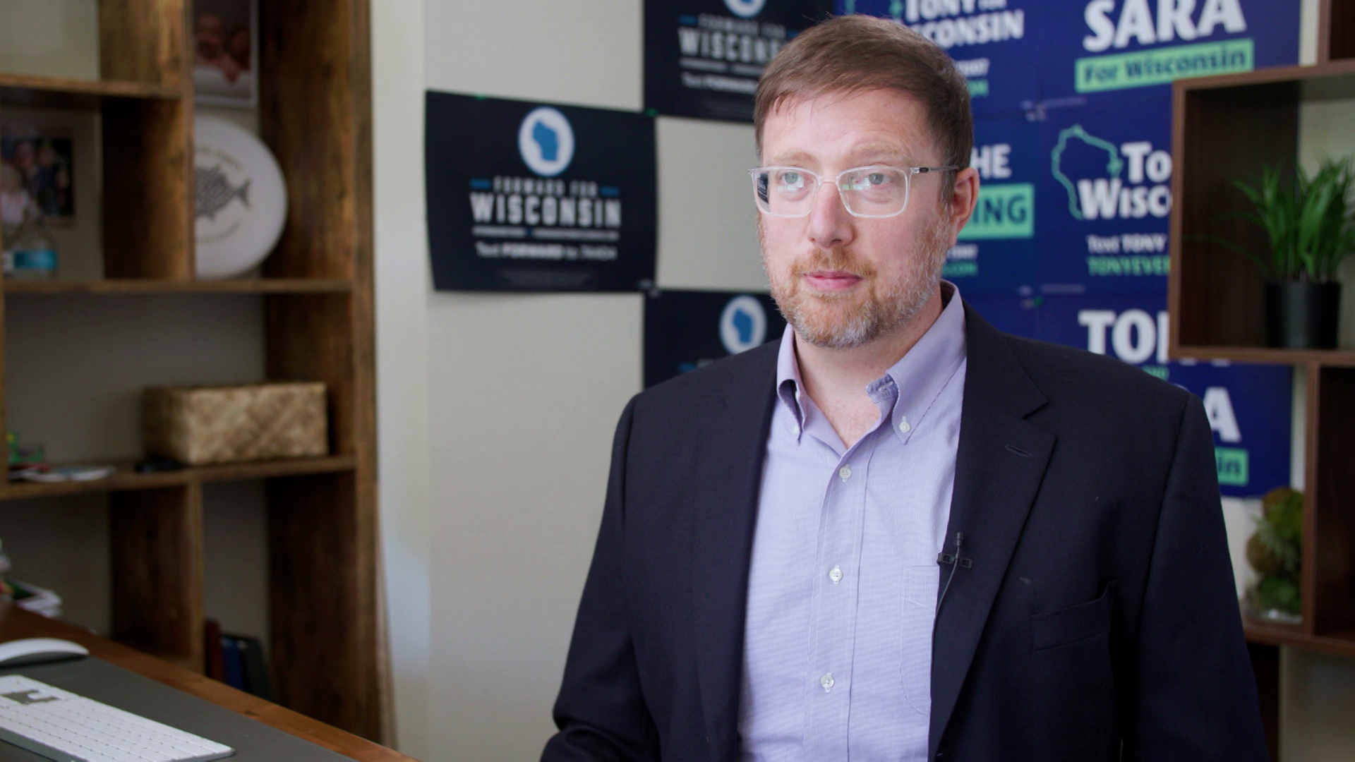 Ben Wikler sits in an office with wooden shelves with plants and other items in the background along with political posters for Democratic Party candidates in the 2022 elections.