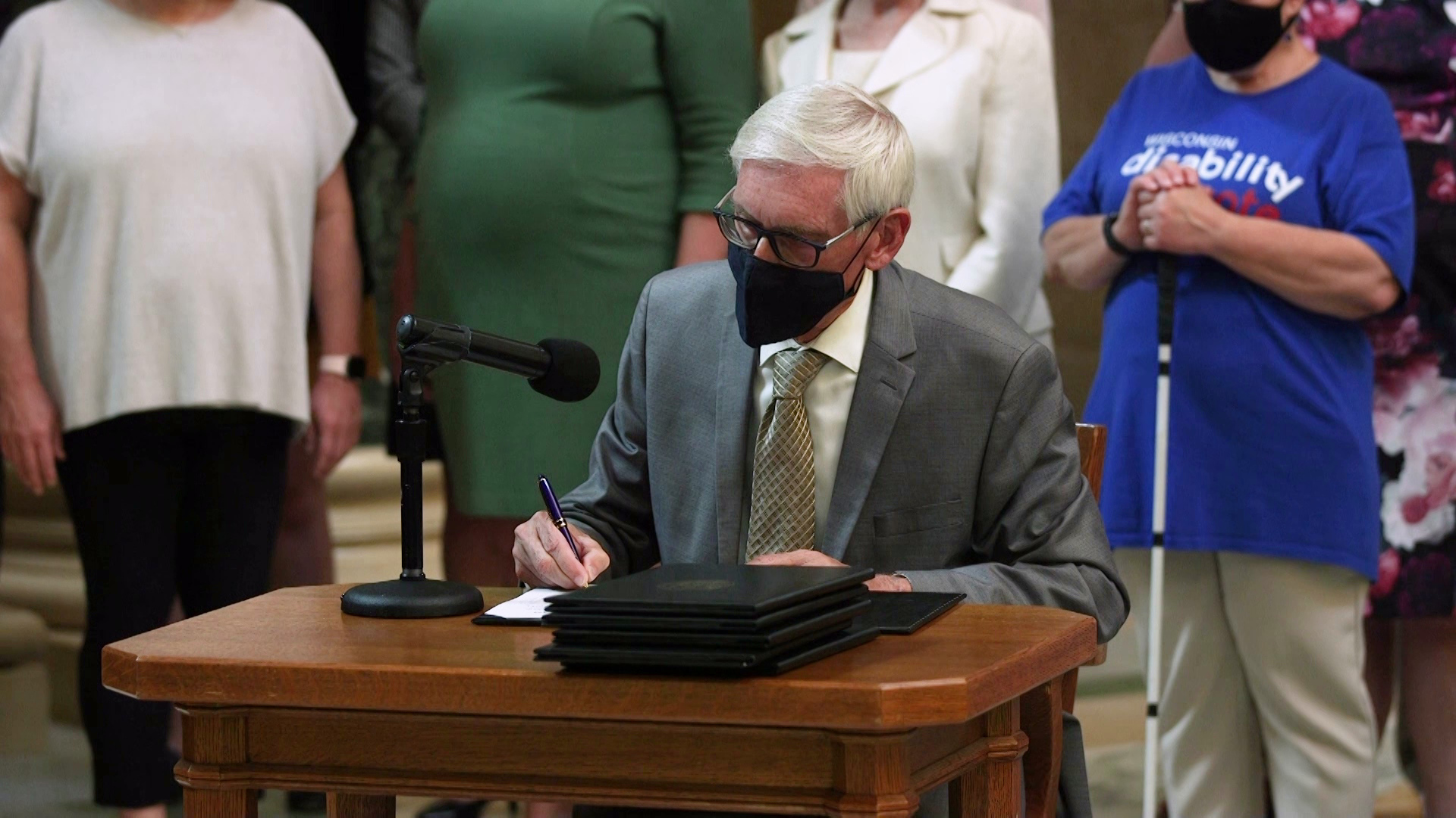 Tony Evers sits at a small, wood table and signs a piece of paper next to a stack of thick folders and a microphone mounted on a stand, with people standing in the background.