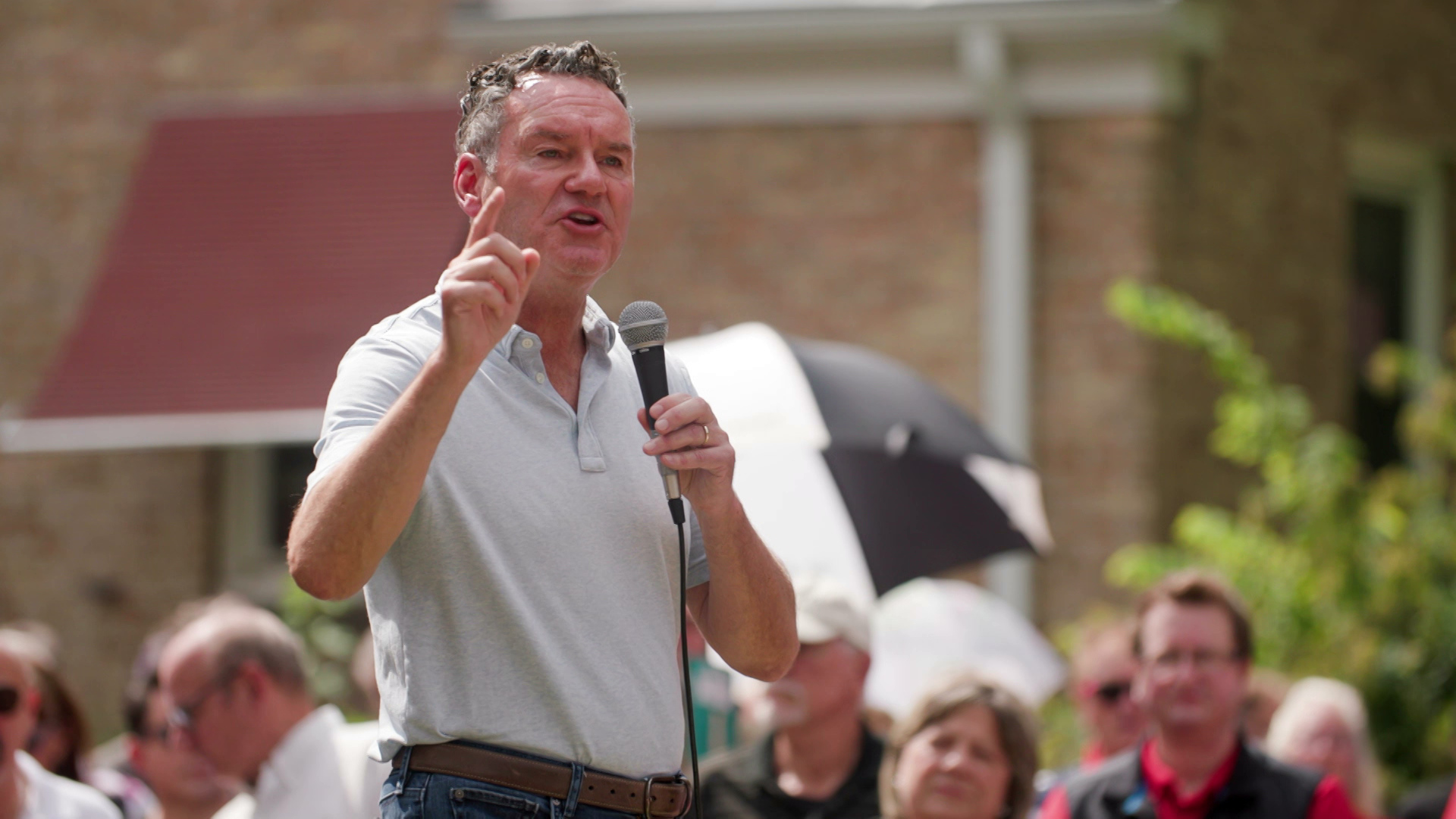 Tim Michels stands and speaks outdoors, holding a microphone in his left hand and gesturing with his right hand, with standing people and a brick building in the background.