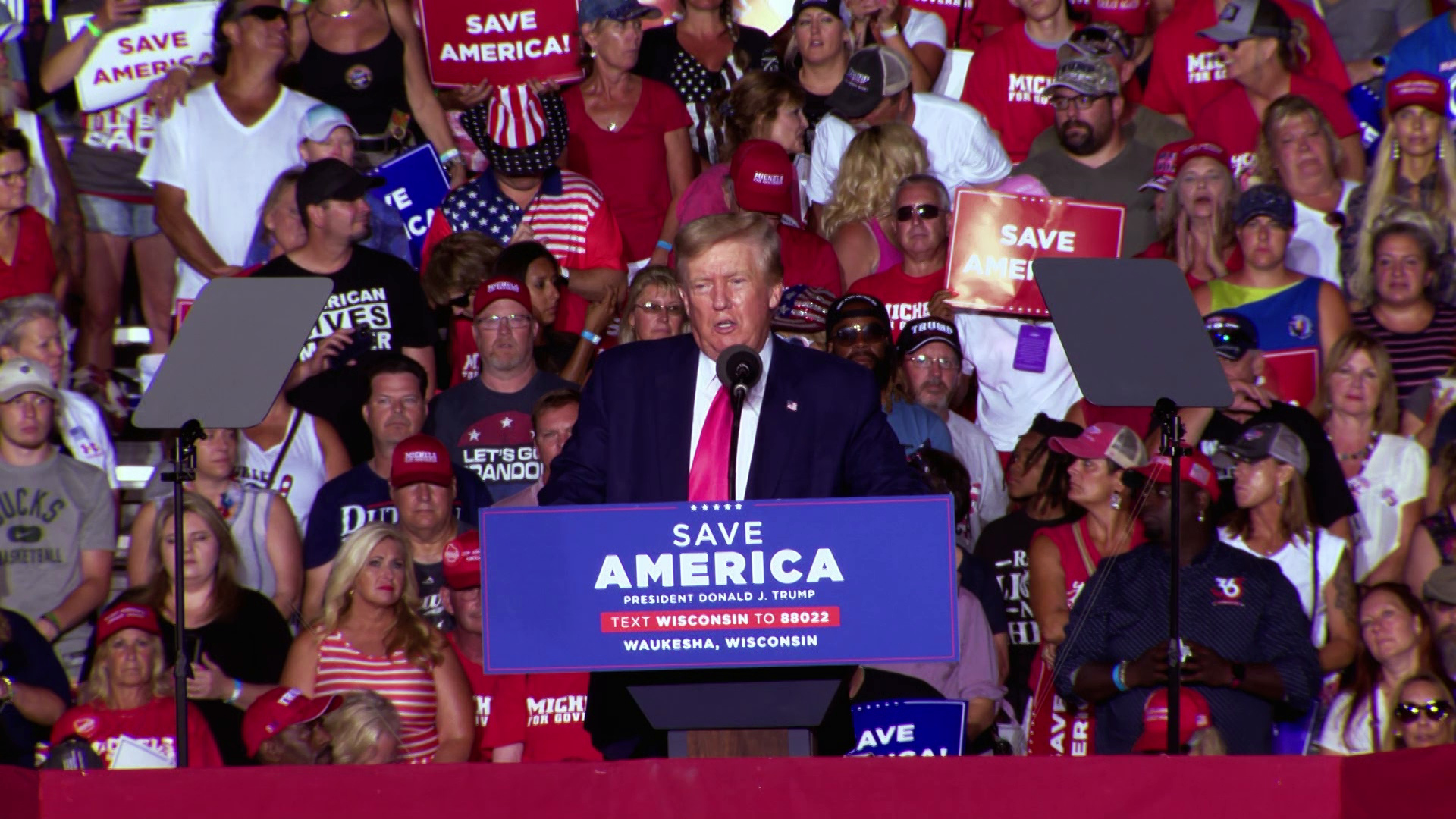 Donald Trump stands behind a podium with a sign reading "Save America" and speaks into a microphone, with teleprompters on either side and an audience of listeners in the background.