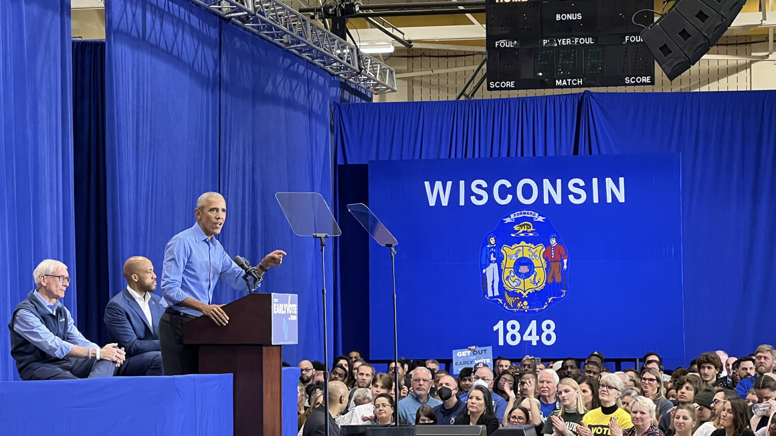 Barack Obama delivers a speech from behind a podium on a stage, with Tony Evers and Mandela Barnes seated behind him, as audience members standing below listen and applaud in a gymnasium with large room-dividing curtains, a Wisconsin flag billboard, and speakers and a scoreboard mounted on the ceiling.