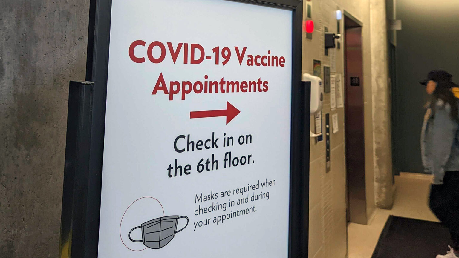 A sign reading COVID-19 Vaccine Appointments with an arrow pointing right, with additional text of Check in on the 6th floor and Masks are required when checking in and during your appointment. stands next to a pair of elevator doors with one person standing in front of them.