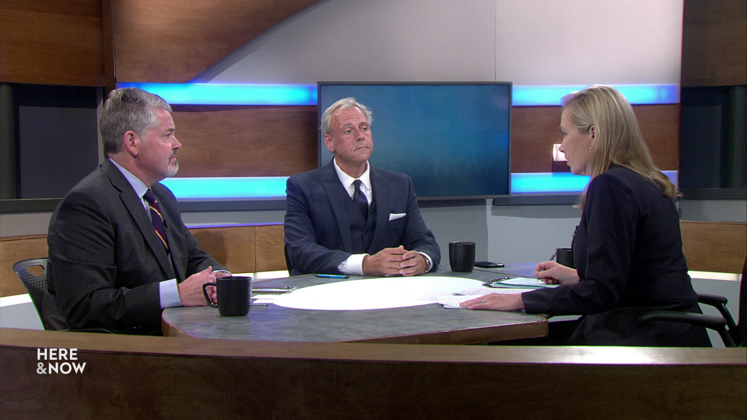 Bill McCoshen and Scot Ross sit next to each other and across from Frederica Freyberg at a desk on the Here & Now set.