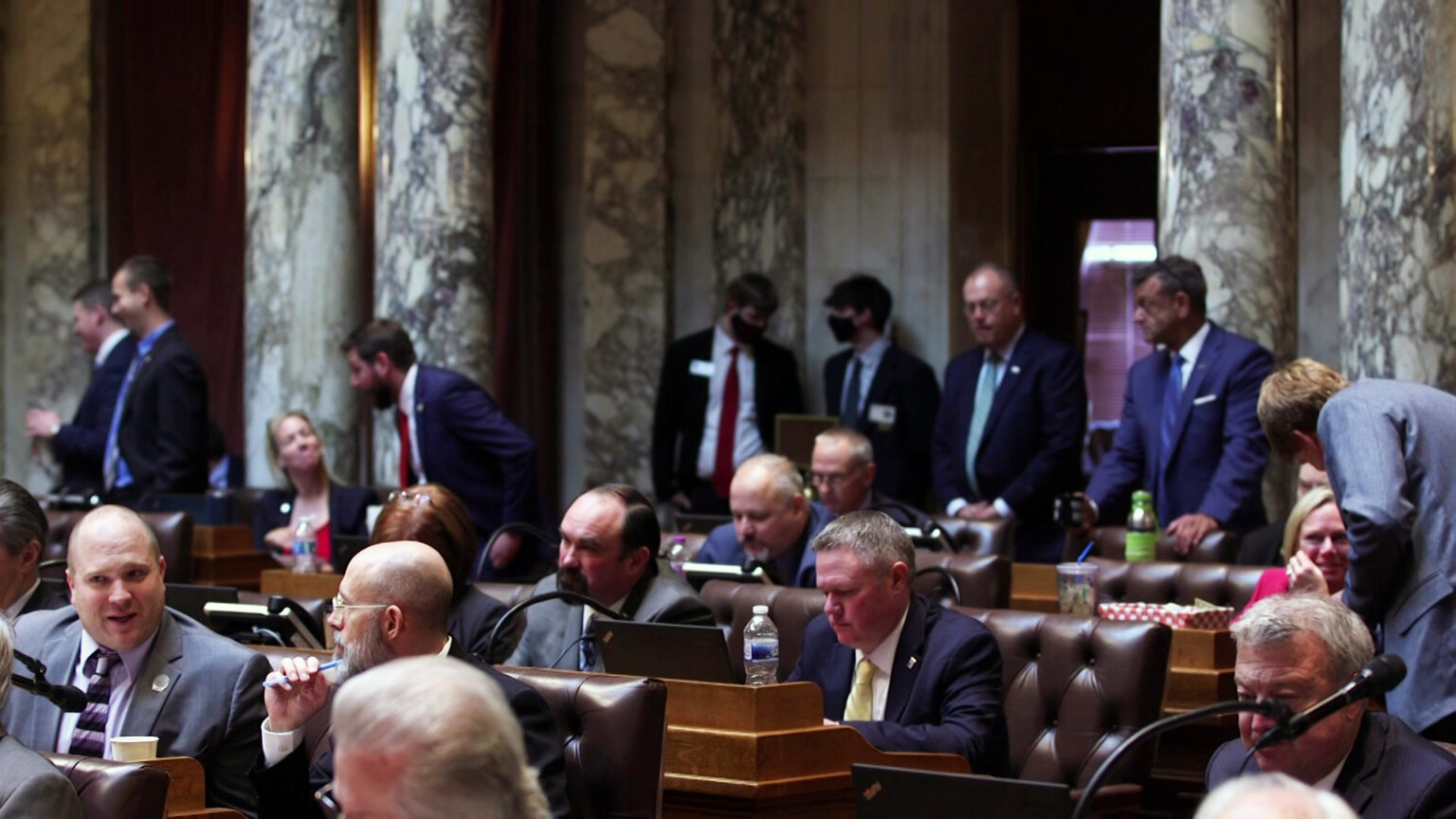 Wisconsin Assembly members and support staff dressed in professional attire sit at desks with large leather-padded chairs or stand in aisles in a room with marble walls and columns.