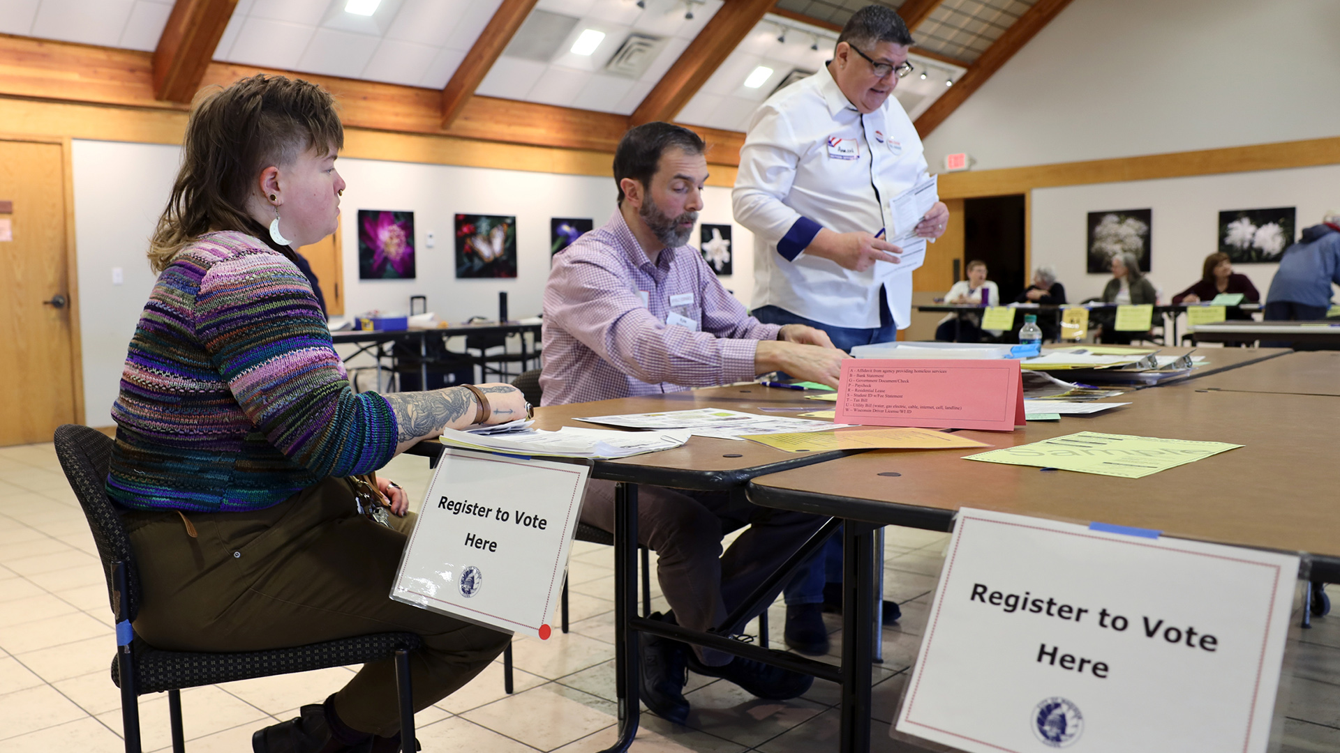 Alix Yarrow sits at a table with Kyle Richmond, with Aaron Schultz standing nearby, with signs reading "Register to Vote Here" affixed to its edge and other people seated at tables in the background of a large room with a pitched ceiling.