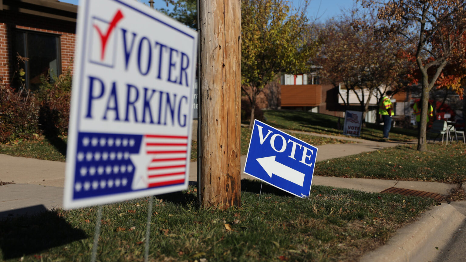 A yard sign reading Vote and with an arrow pointing left stands in turf between a sidewalk and road, with an out-of-focus sign reading Voter Parking in the foreground and with trees and buildings in the background.