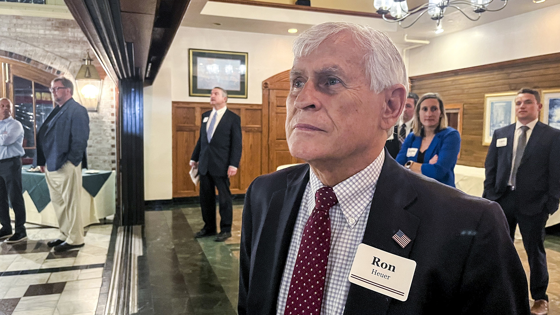 Ron Heuer stands while wearing a U.S. flag lapel pin and nametag reading "Ron Heuer" in a room with marble-patterned flooring and segments of wood-paneled walls, with other people in business attire standing in the background.