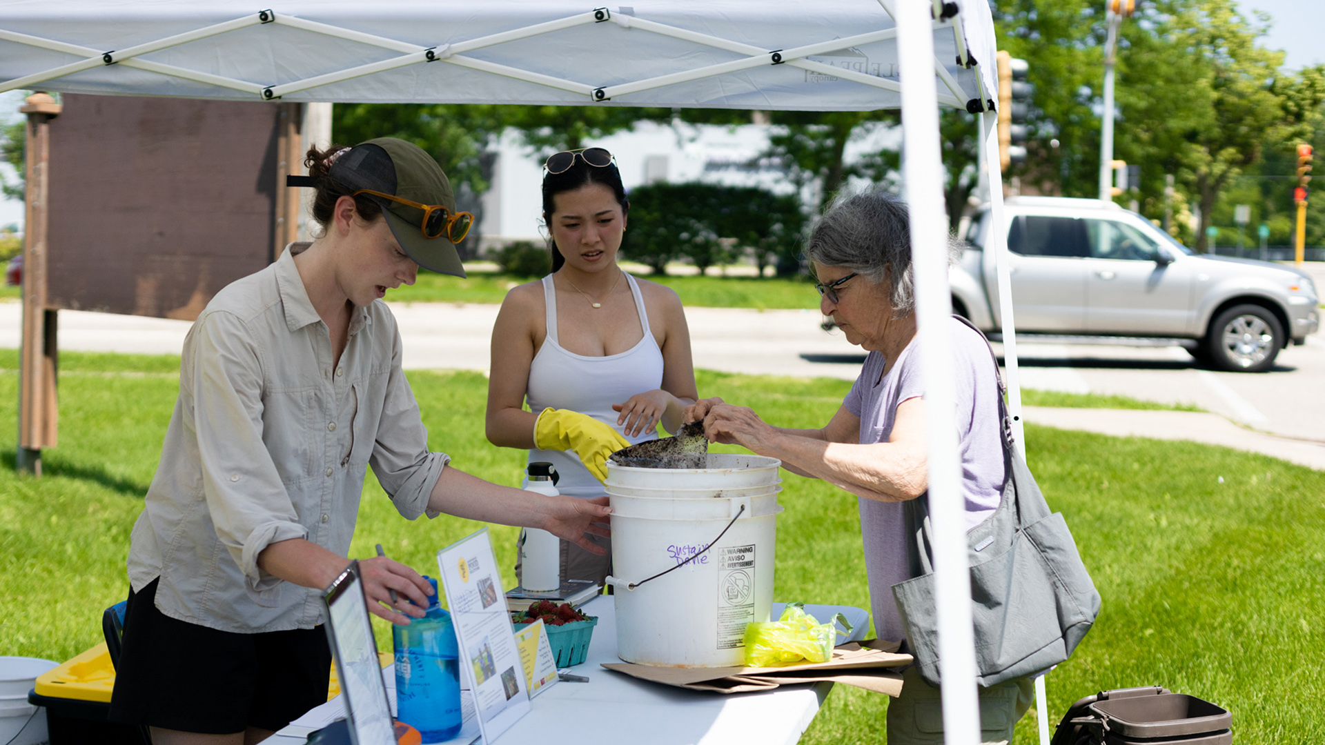 Three people stand around a table standing on a lawn under an outdoor canopy tent, with two helping the third empty food scraps into a five-gallon plastic bucket.