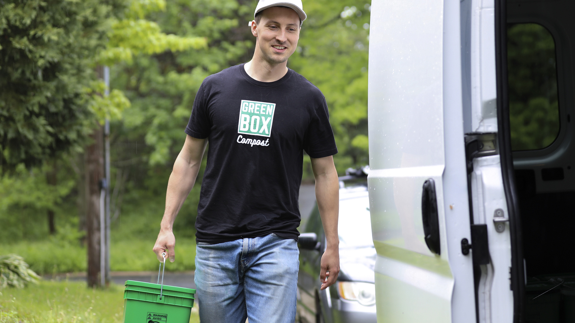 Ben Stanger carries a plastic bucket with a closed top toward the open sliding door of a cargo van, with trees and a parked car in the background.